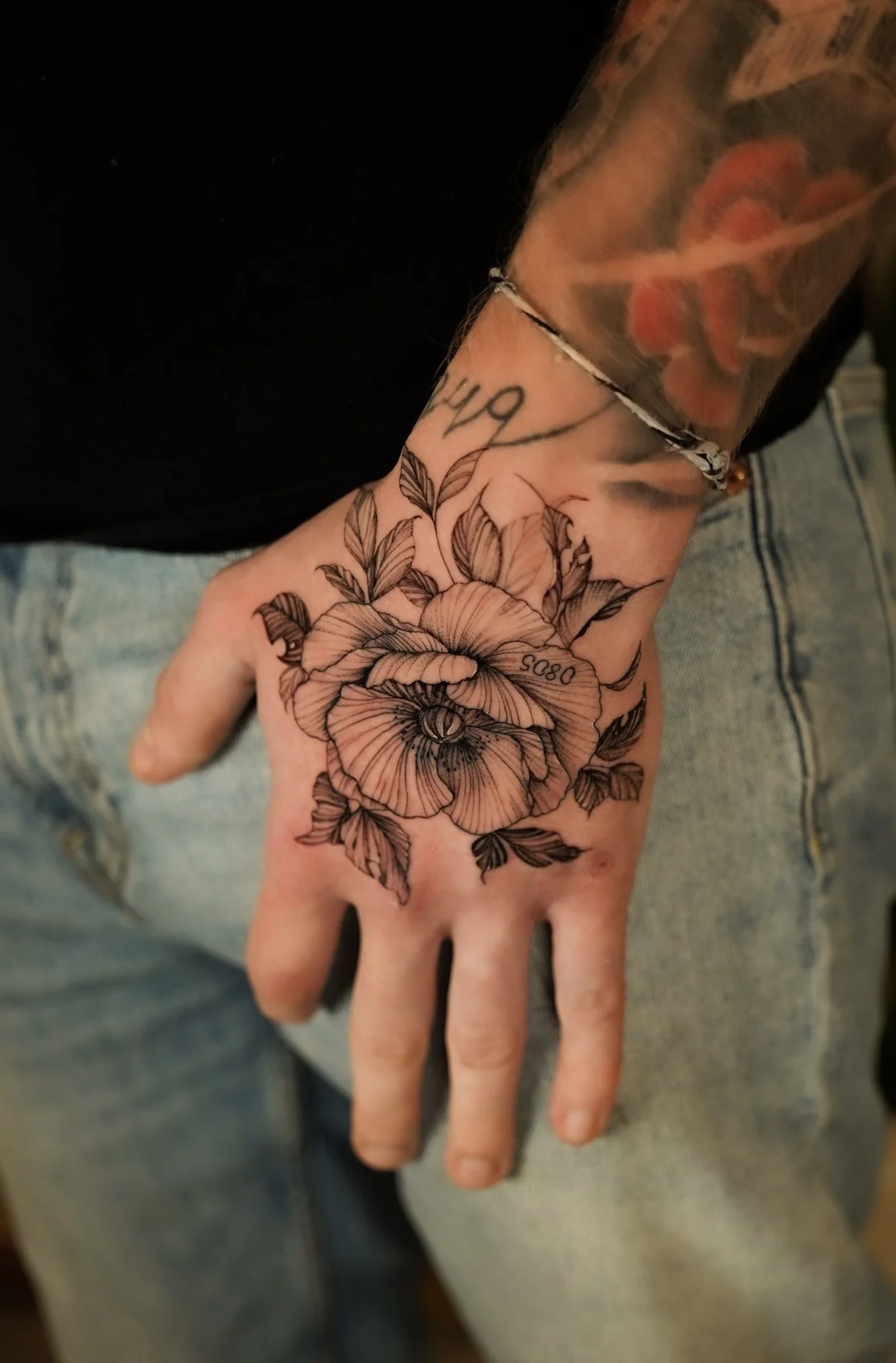 A close-up of a person's hand with a detailed black ink floral tattoo featuring roses and leaves. The person is wearing a black shirt, a silver bracelet, and light blue jeans.