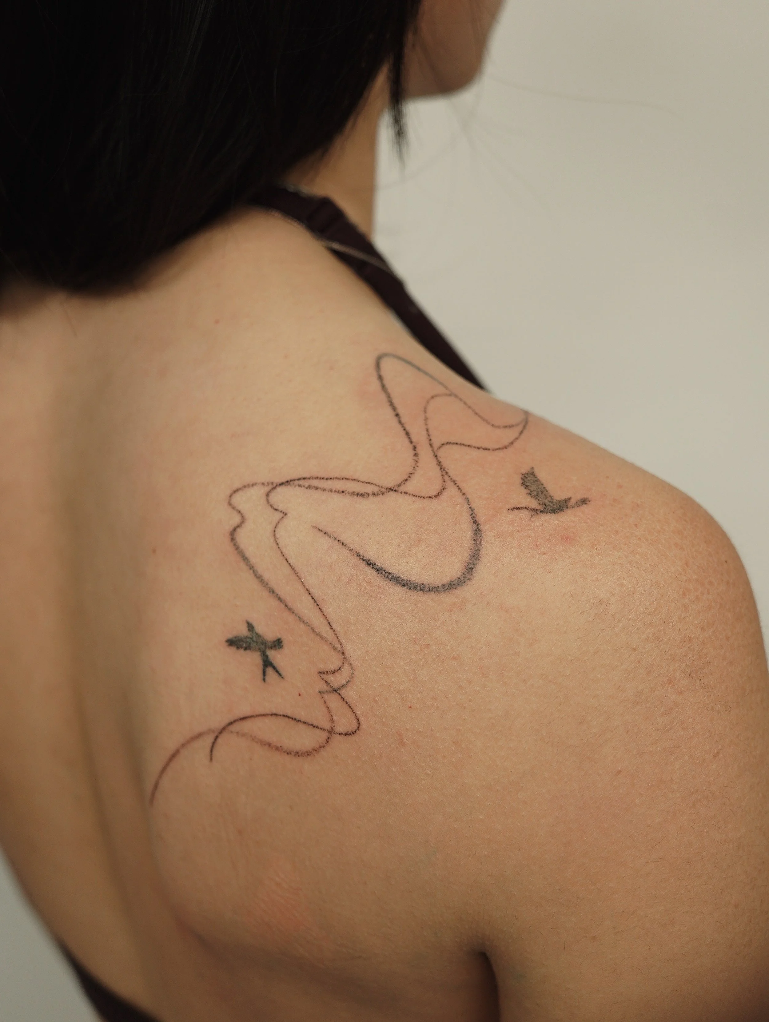 A close-up of a woman's shoulder with a tattoo of a continuous line drawing of a bird and two smaller flying birds, with her dark hair partially visible.