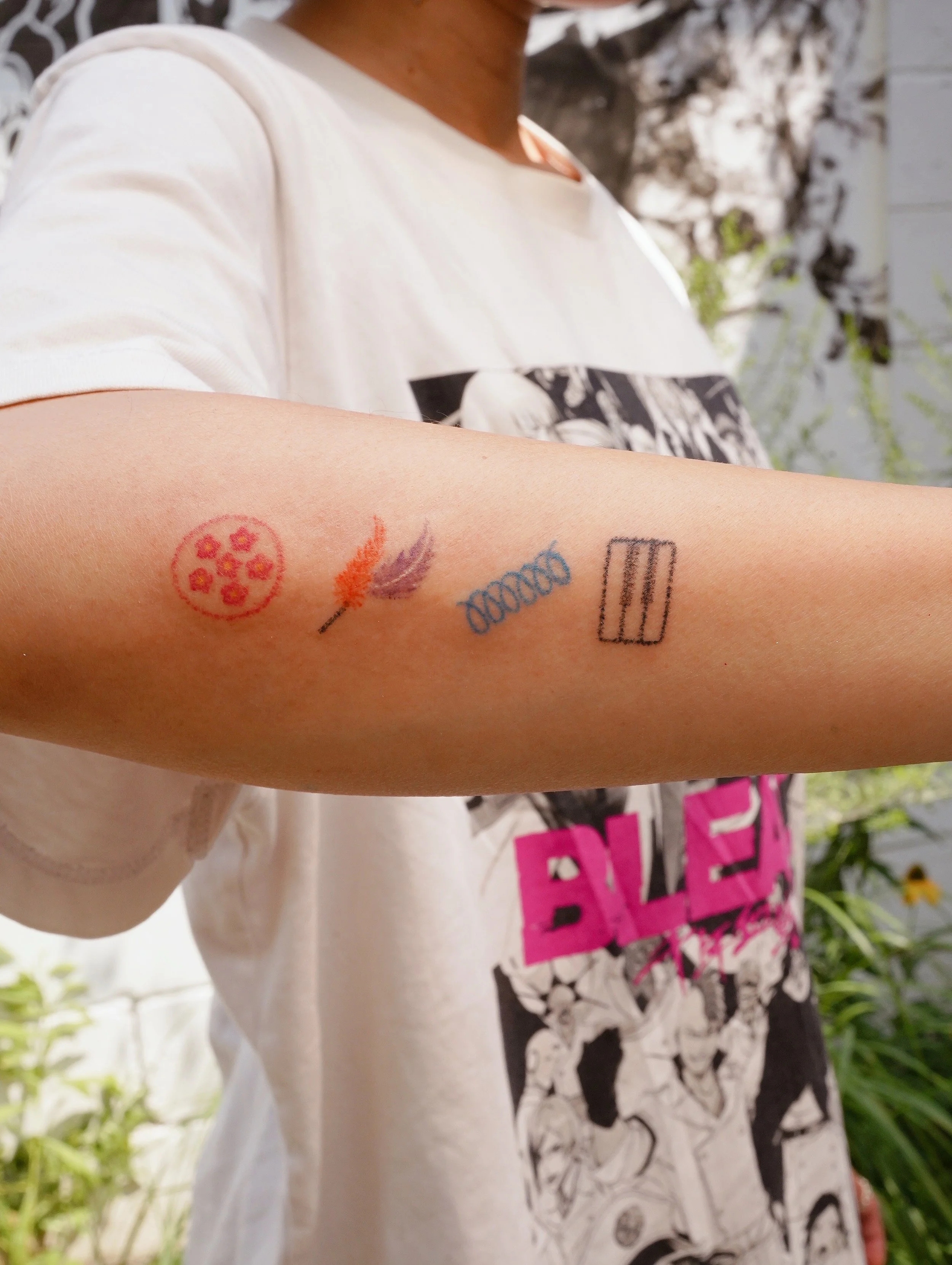 A person's arm with five tattoos: a flower circle, a feather, a double helix, a barcode, and a small triangular shape.