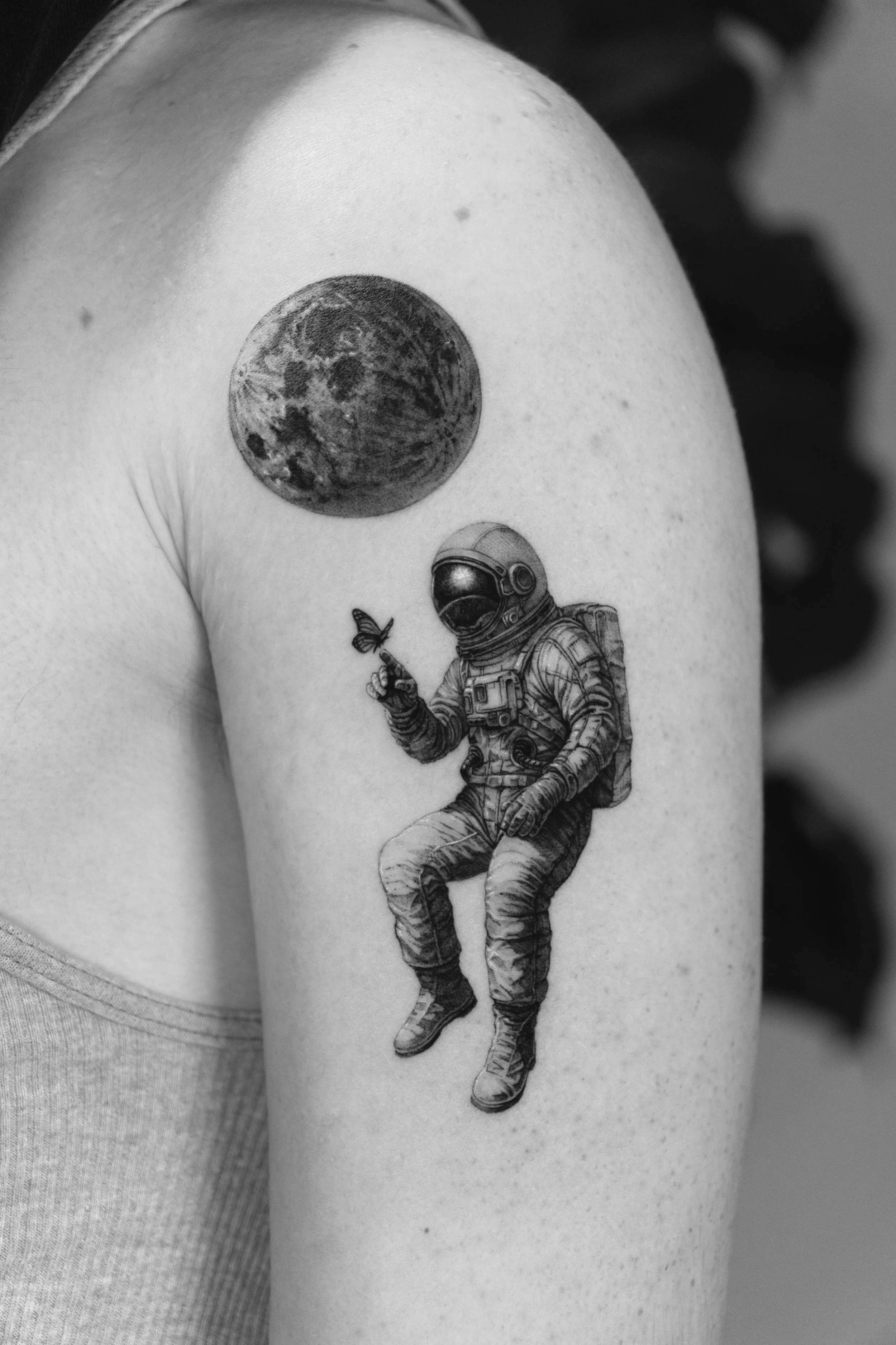 Tattoo of an astronaut sitting, holding a butterfly on its index finger, with a planet in the background on a person's upper arm.