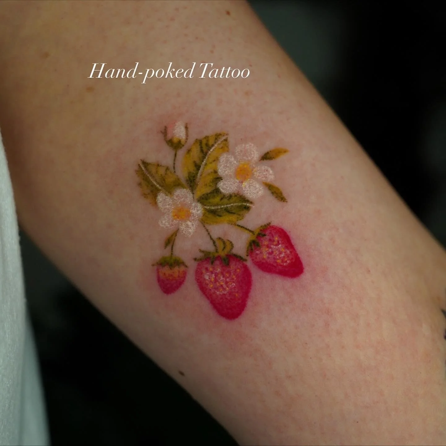 Temporary tattoo of strawberries, leaves, and white flowers with yellow centers on skin, labeled 'Hand-poked Tattoo'