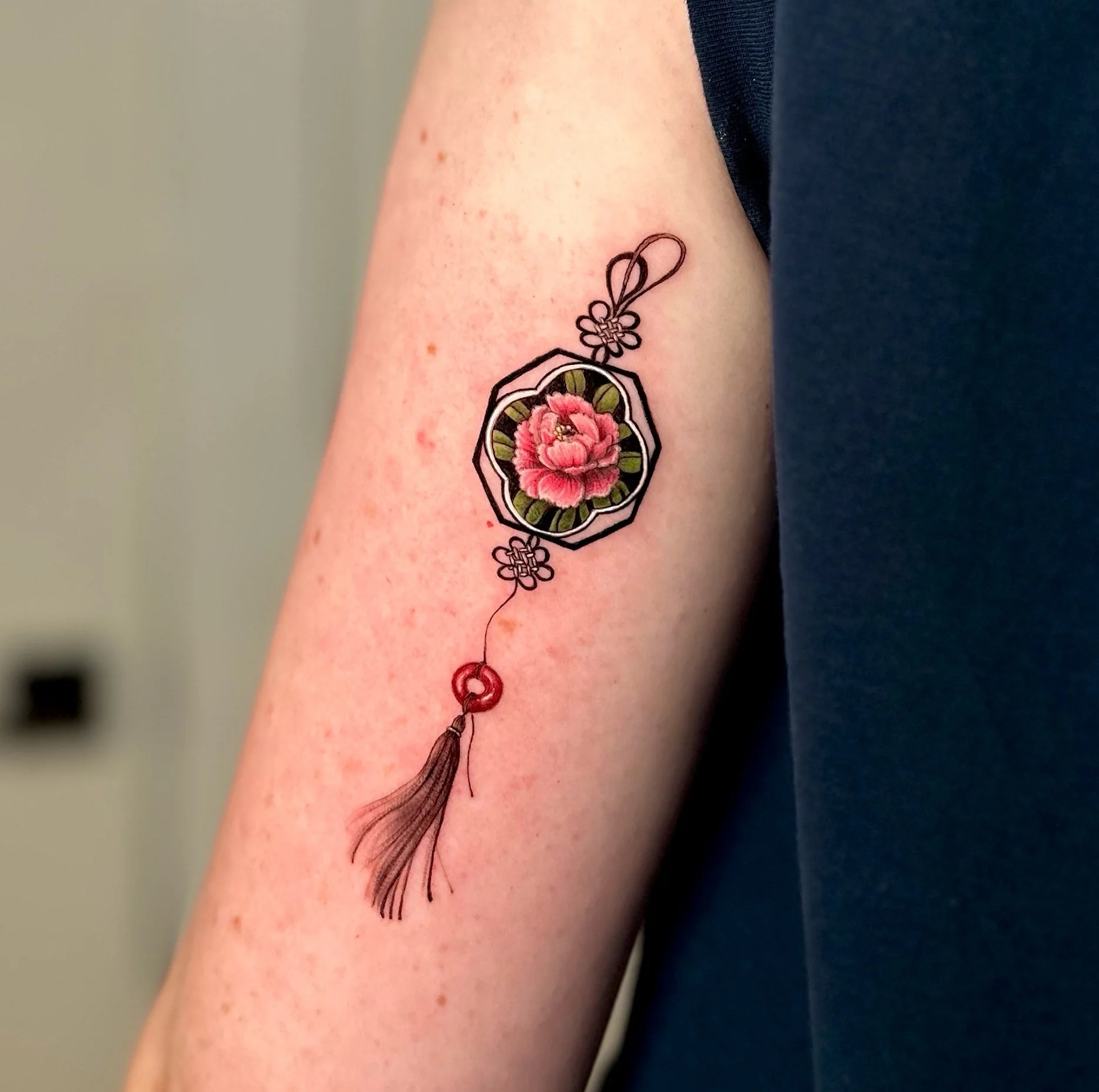 Colorful tattoo on person's upper arm featuring a stained-glass style pink flower, black outlines, and decorative elements including a tassel or charm.