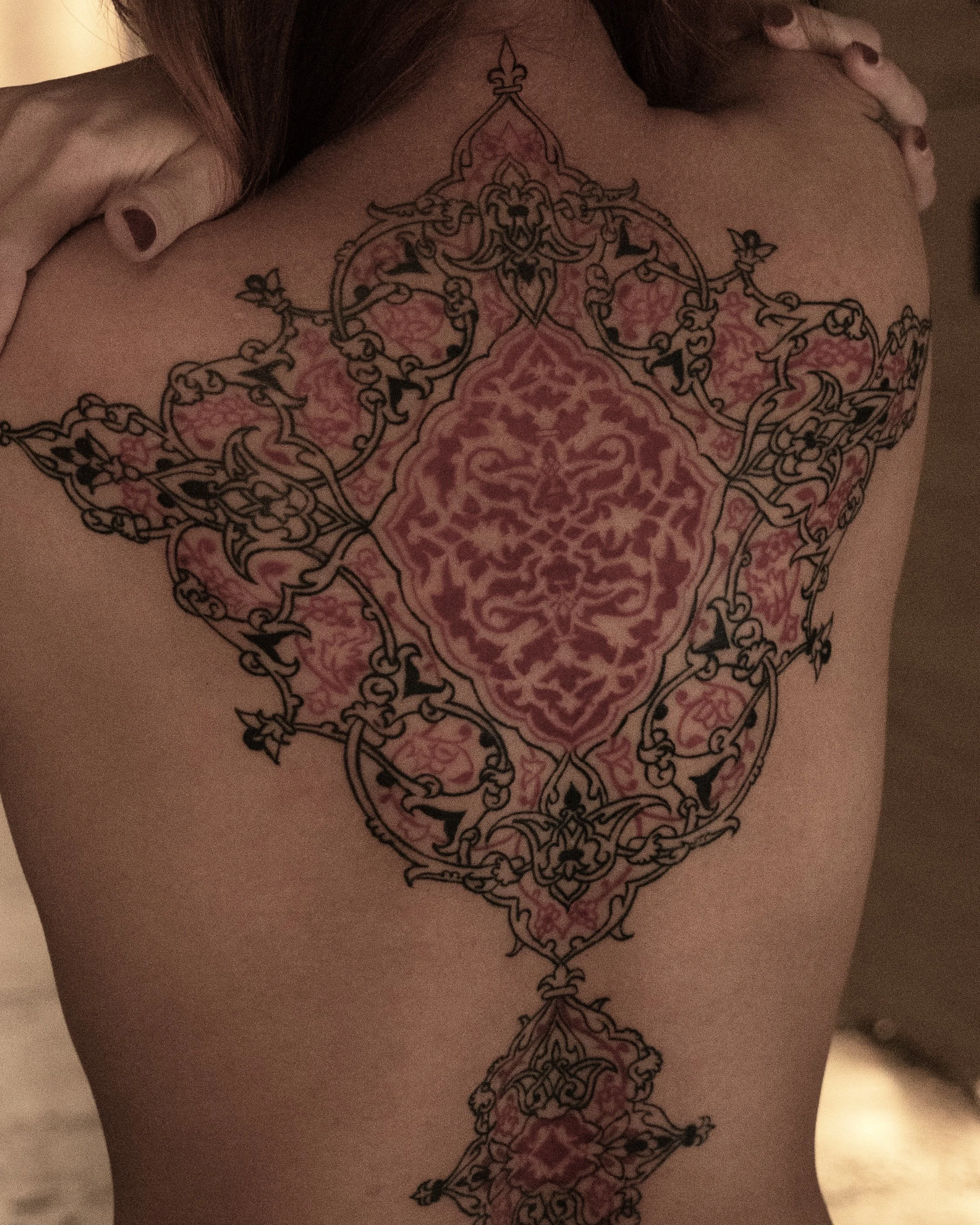 A person's back with an intricate, symmetric tattoo design featuring floral and ornamental patterns in black ink, with pink shading highlighting the patterns.