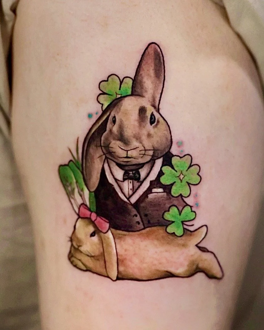 Tattoo of a cartoon rabbit dressed in a tuxedo with a bowtie, surrounded by shamrocks and a second bunny wearing a pink bow.