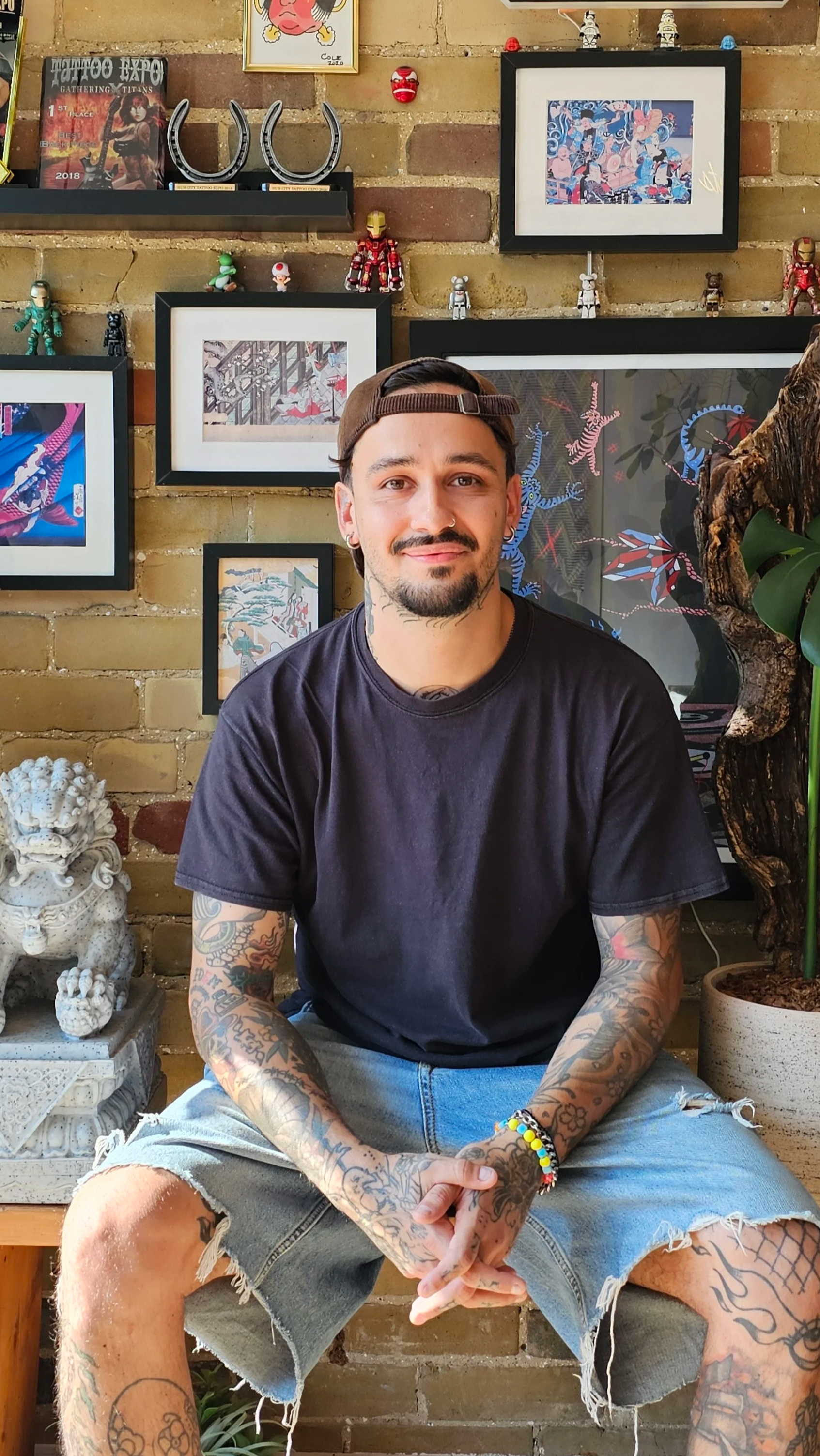 A young man with tattoos sitting in a room with artwork on the brick wall behind him, including framed pictures, small figurines, and decorative pieces.