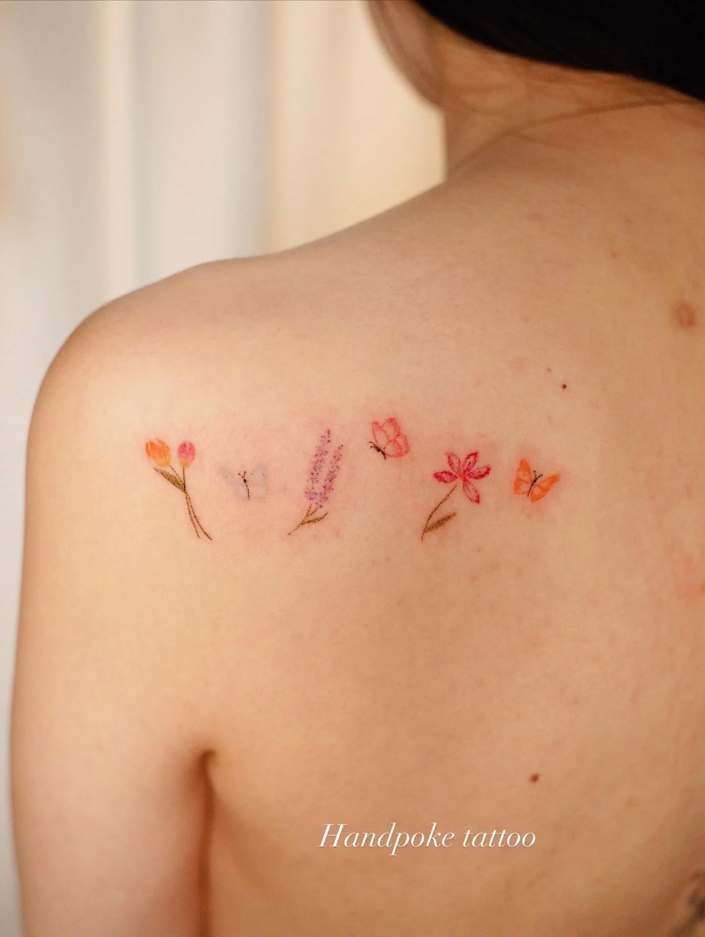 A person's shoulder with a small tattoo of various colorful flowers and butterflies. The tattoo is labeled 'Handpoke tattoo'.