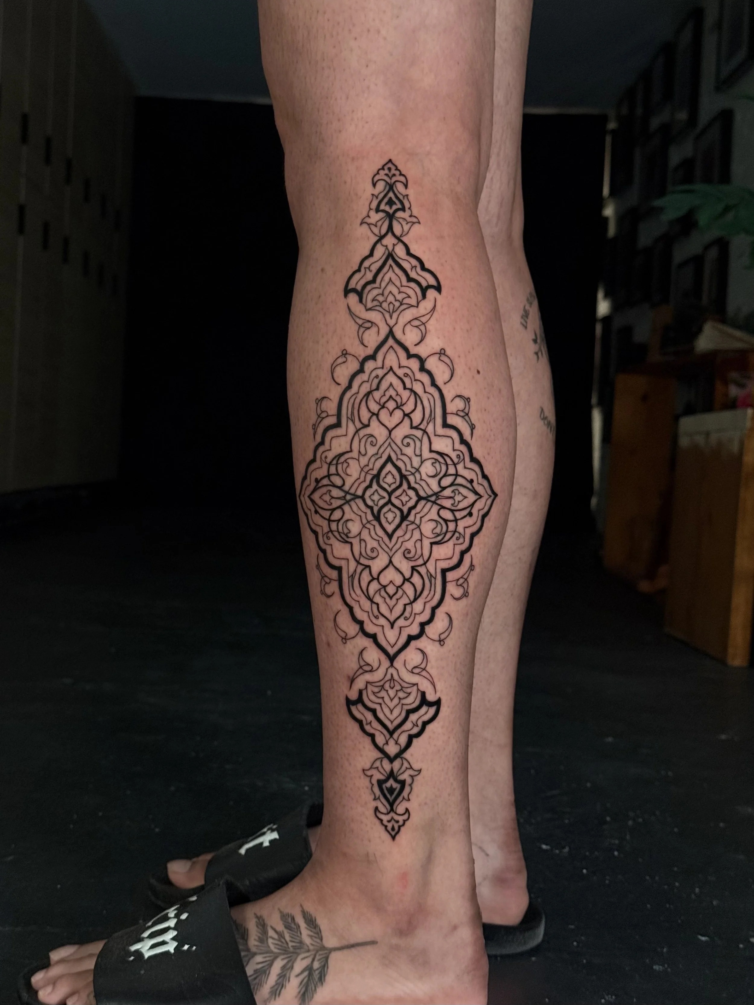 A person's lower leg with a large intricate black tattoo in a symmetrical geometric pattern.