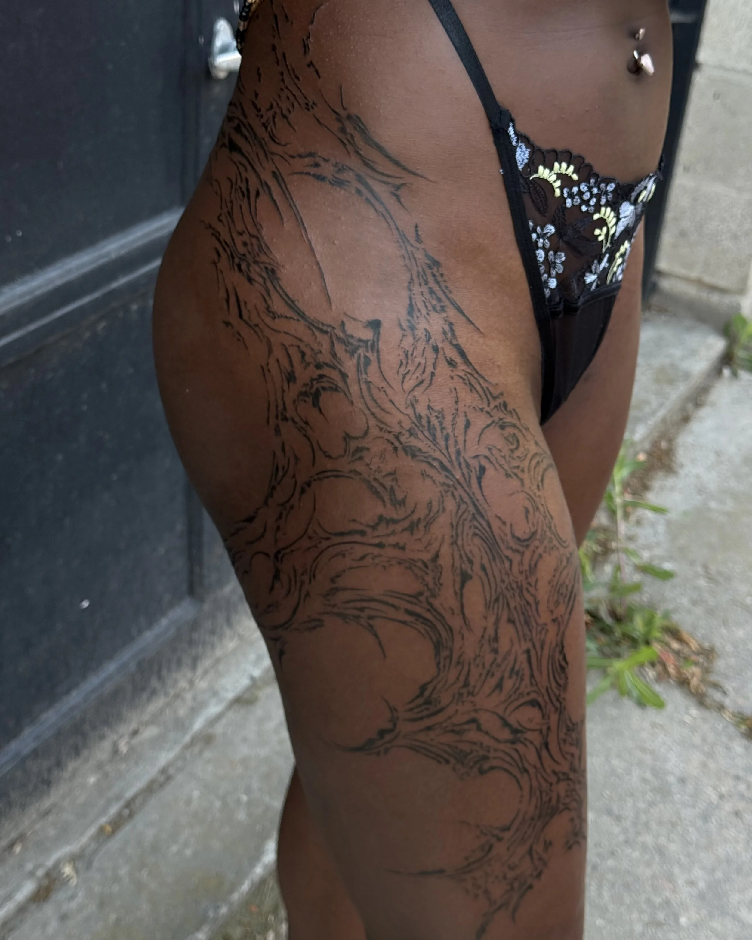 Close-up of a person's thigh with black tattoo art resembling swirling patterns, wearing black lace underwear with floral embroidery, outdoors on concrete and grass.