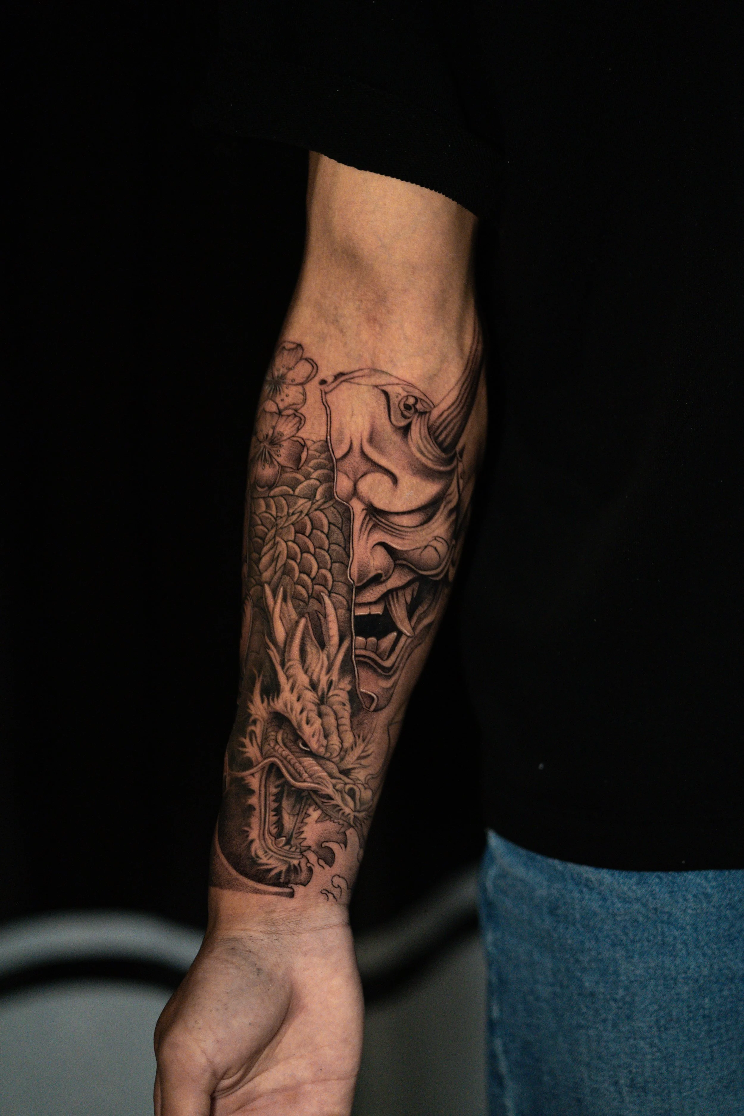 Tattoo of a traditional Asian dragon and a smiling Kabuki-style face on a person's arm.