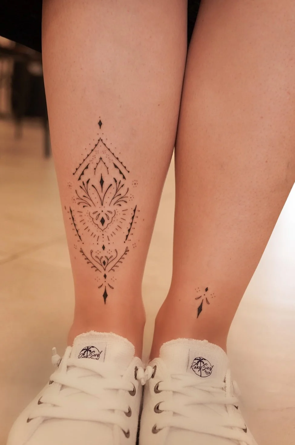 Two tattoos on person's legs, one larger on the left shin with intricate geometric and floral design, and a small, simple black ink design on the right ankle.
