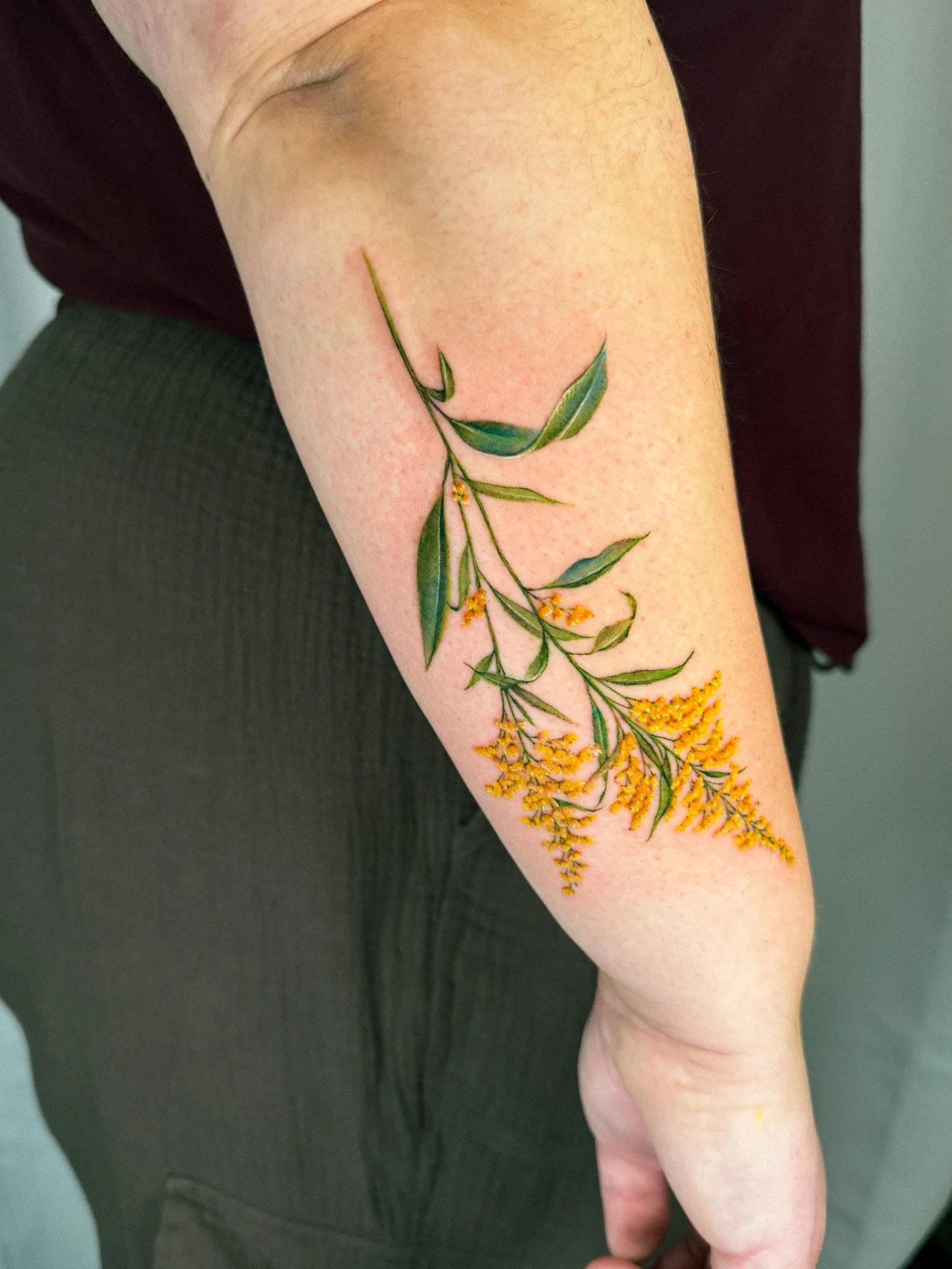 A person's forearm with a detailed botanical tattoo of green leaves and yellow flowers.