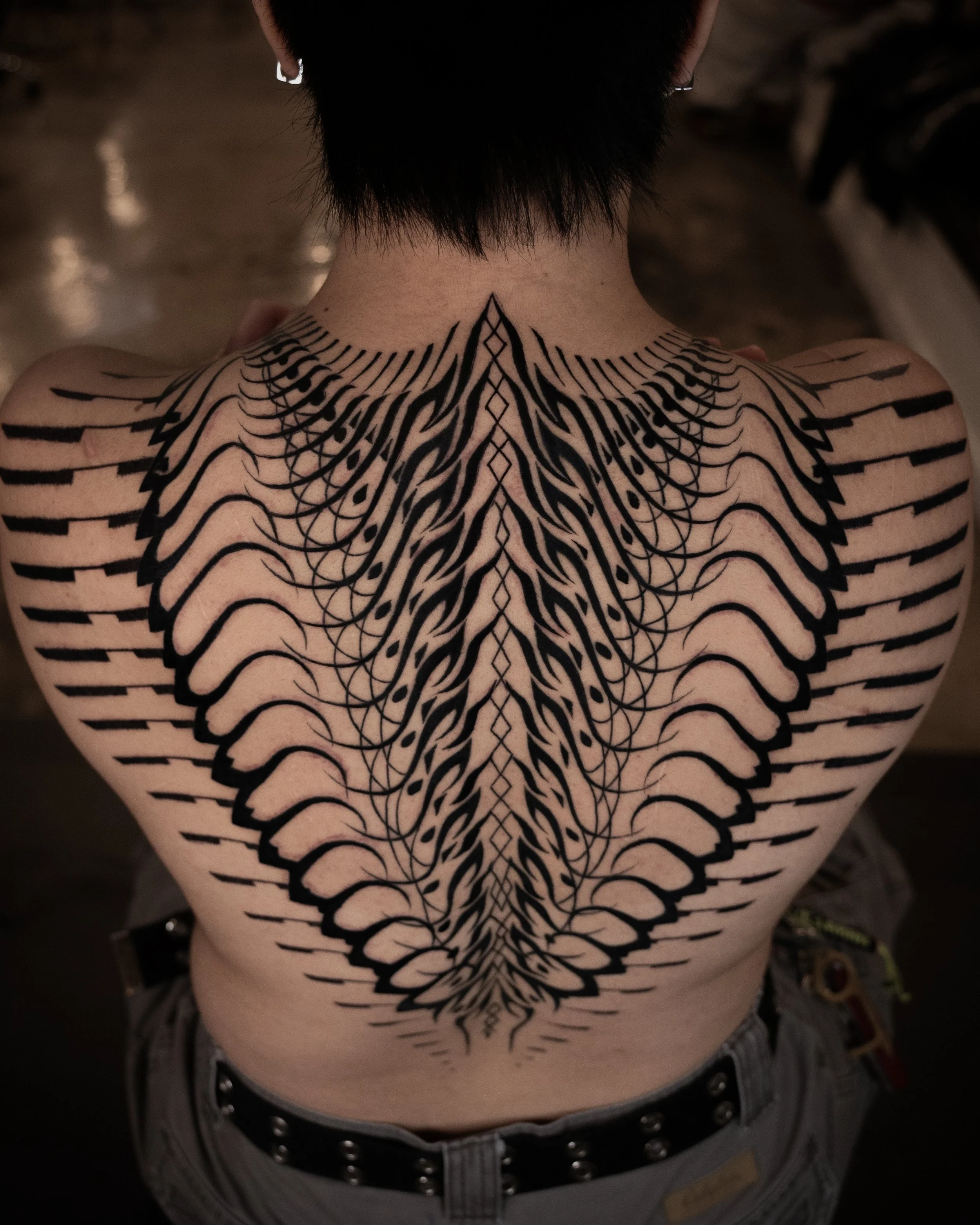 Back of a person with short dark hair, wearing earrings, and a large black tribal-style tattoo on their back and shoulders.
