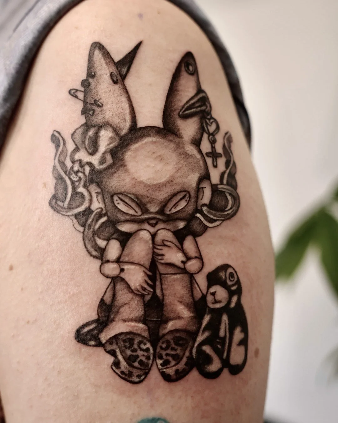 Black and gray tattoo of a cartoonish character with animal ears, coffee beans for eyes, and wearing casual clothing, holding its chest. The character has a cat face mask on the ground, a teddy bear, and various symbols like a cross and a fish on its