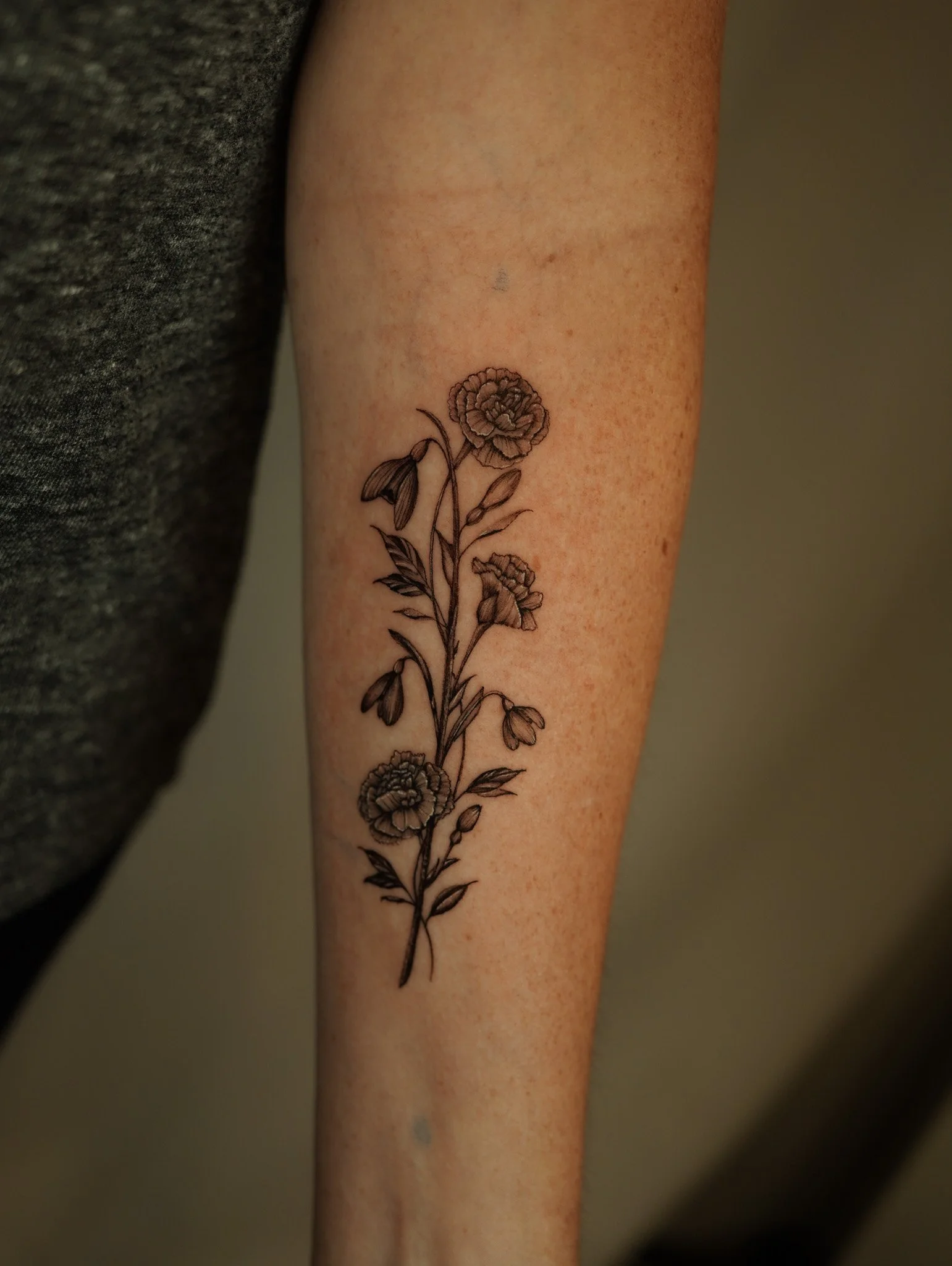A detailed black ink floral tattoo on a person's forearm, featuring multiple flowers, leaves, and buds.
