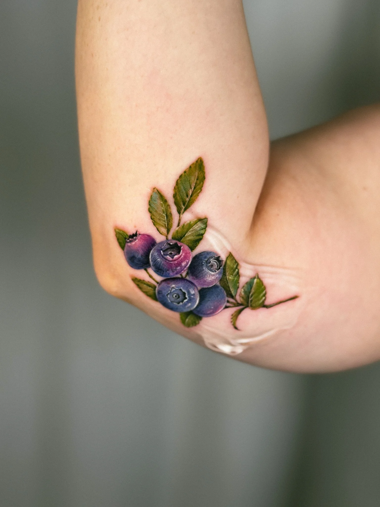Tattoo of blueberries and green leaves on person's inner elbow.