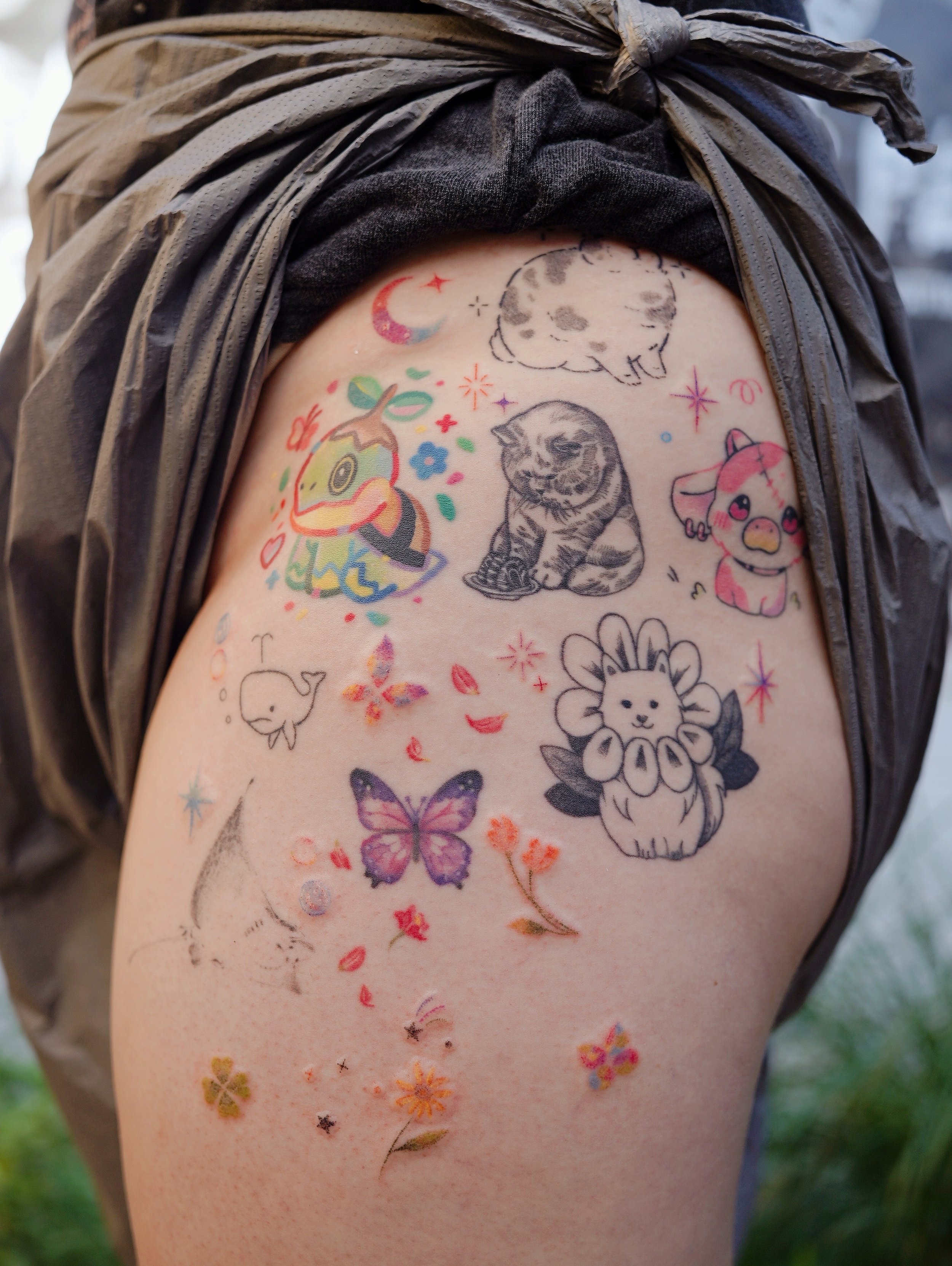 Collection of colorful temporary tattoos on upper arm, including a whale, butterfly, lion, cat, unicorn, moon, cloud, flower, and various small decorative elements.