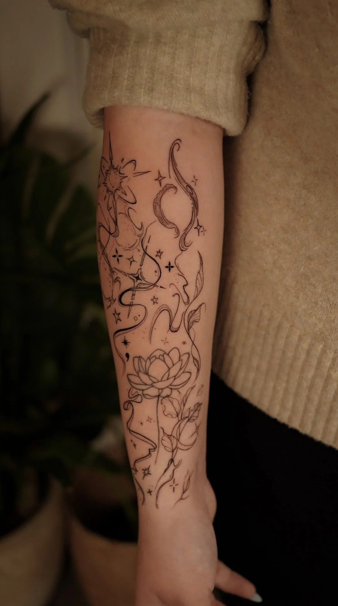 Line art tattoo of a star, swirling smoke, a lotus flower, and celestial stars on a person's forearm.