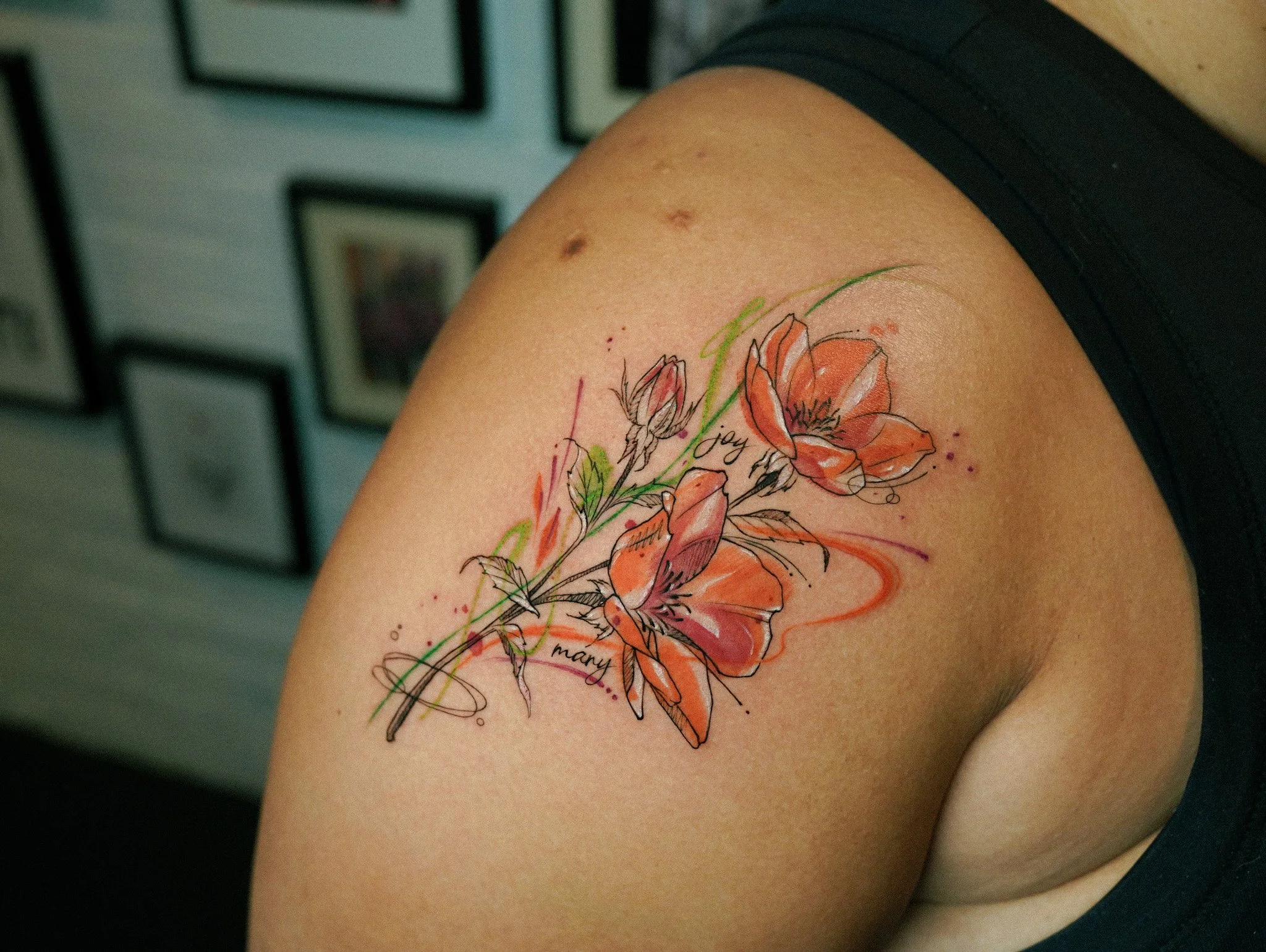 A colorful floral tattoo featuring orange flowers, green accents, and black ink details on a person's shoulder and upper arm.