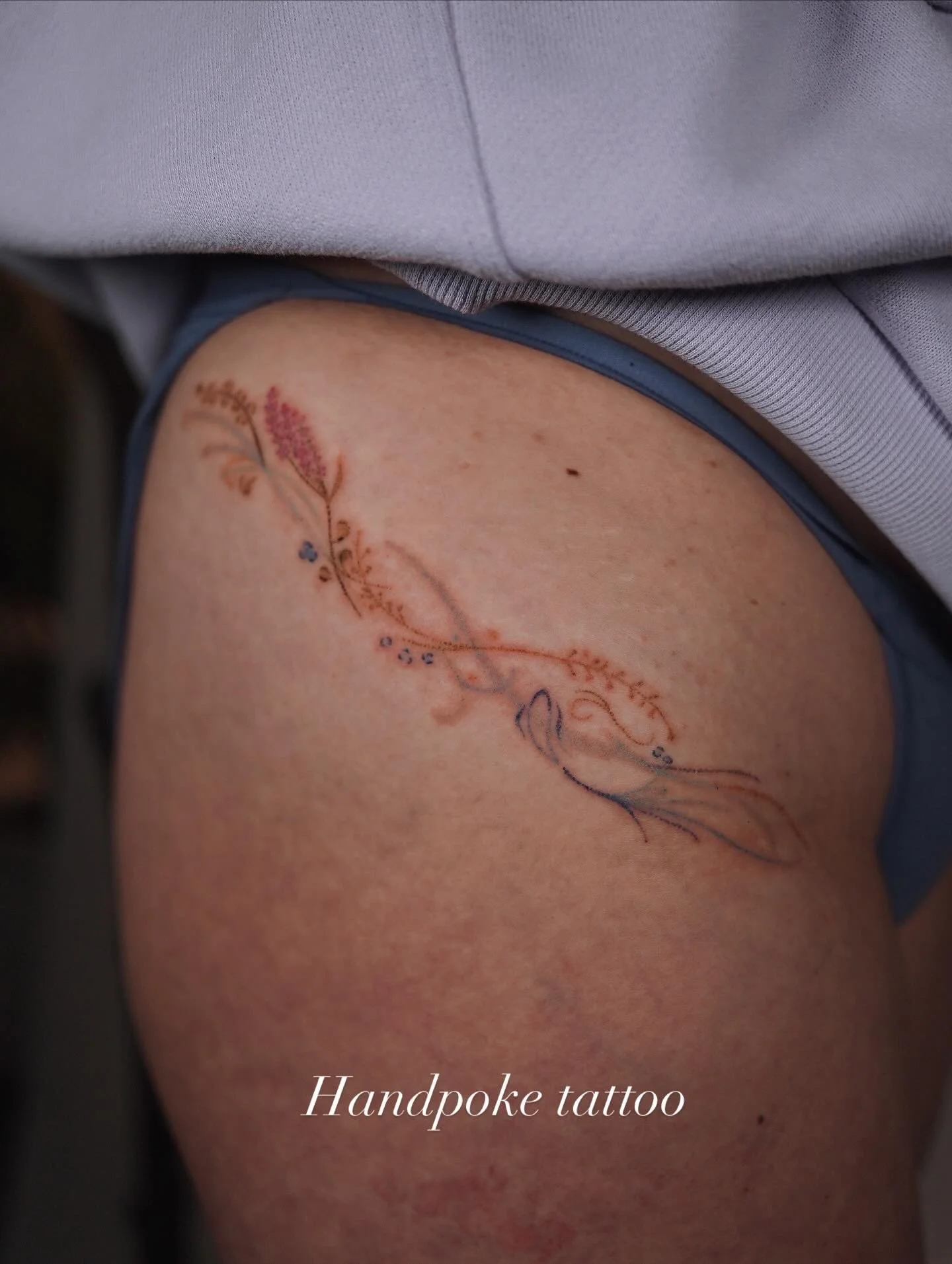 A tattoo of a floral design with a hummingbird on a person's upper arm or shoulder, featuring pink and blue flowers and the text 'Handpoke tattoo' at the bottom.