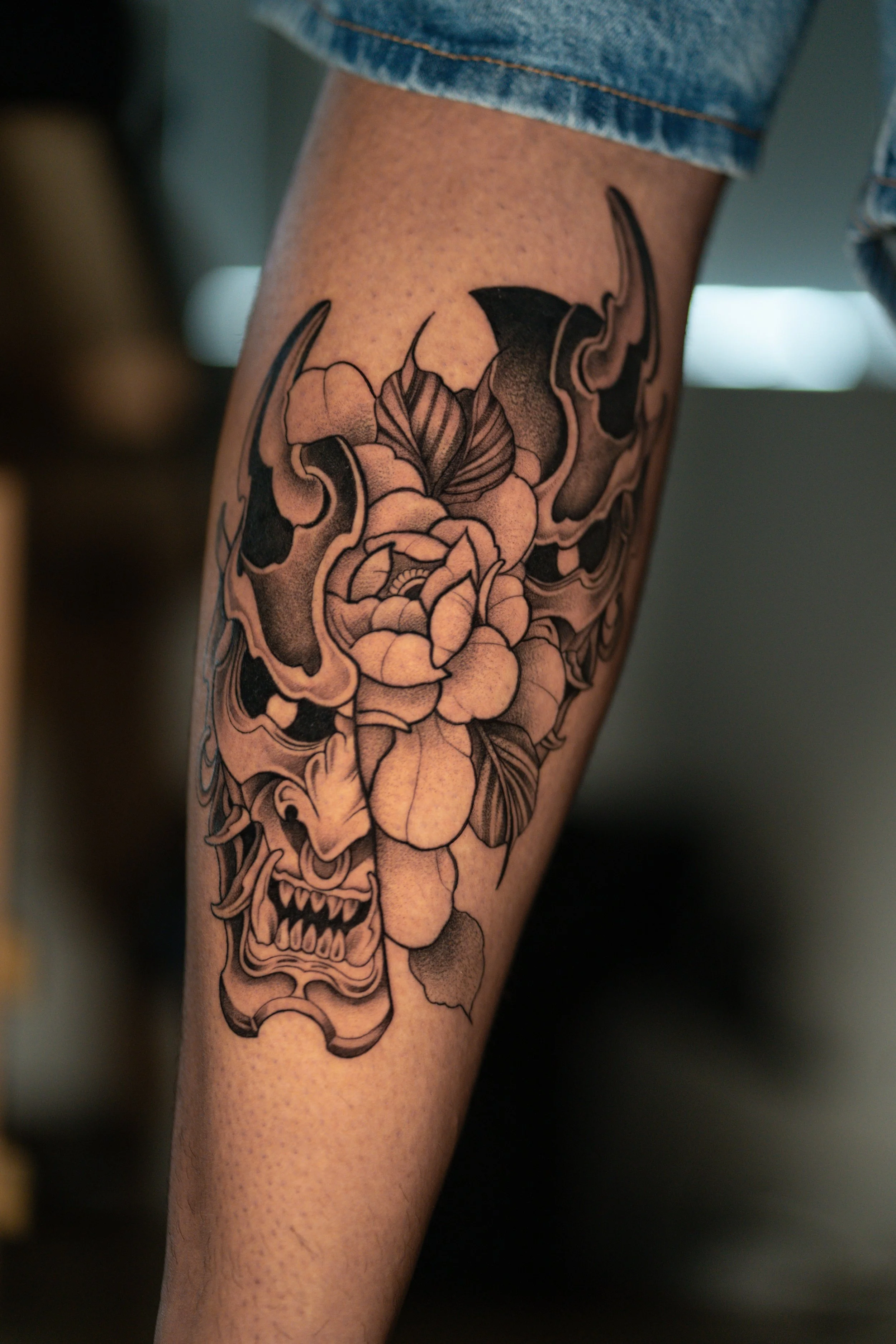 Black and gray tattoo on person's forearm depicting a fierce Asian-style demon mask with sharp teeth, prominent brow, and detailed floral elements surrounding it.