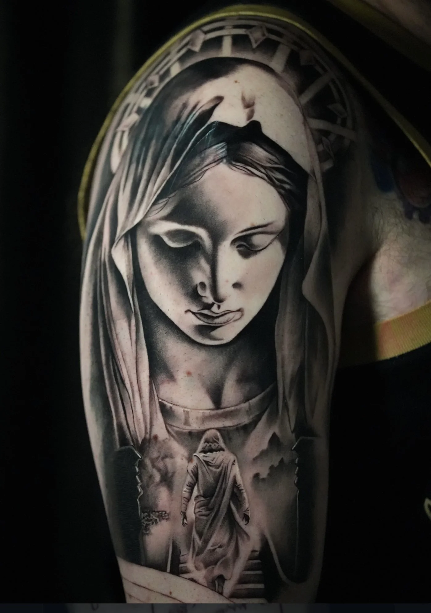 Black and gray tattoo of a woman's face with a halo and a figure in a robe walking down stairs.