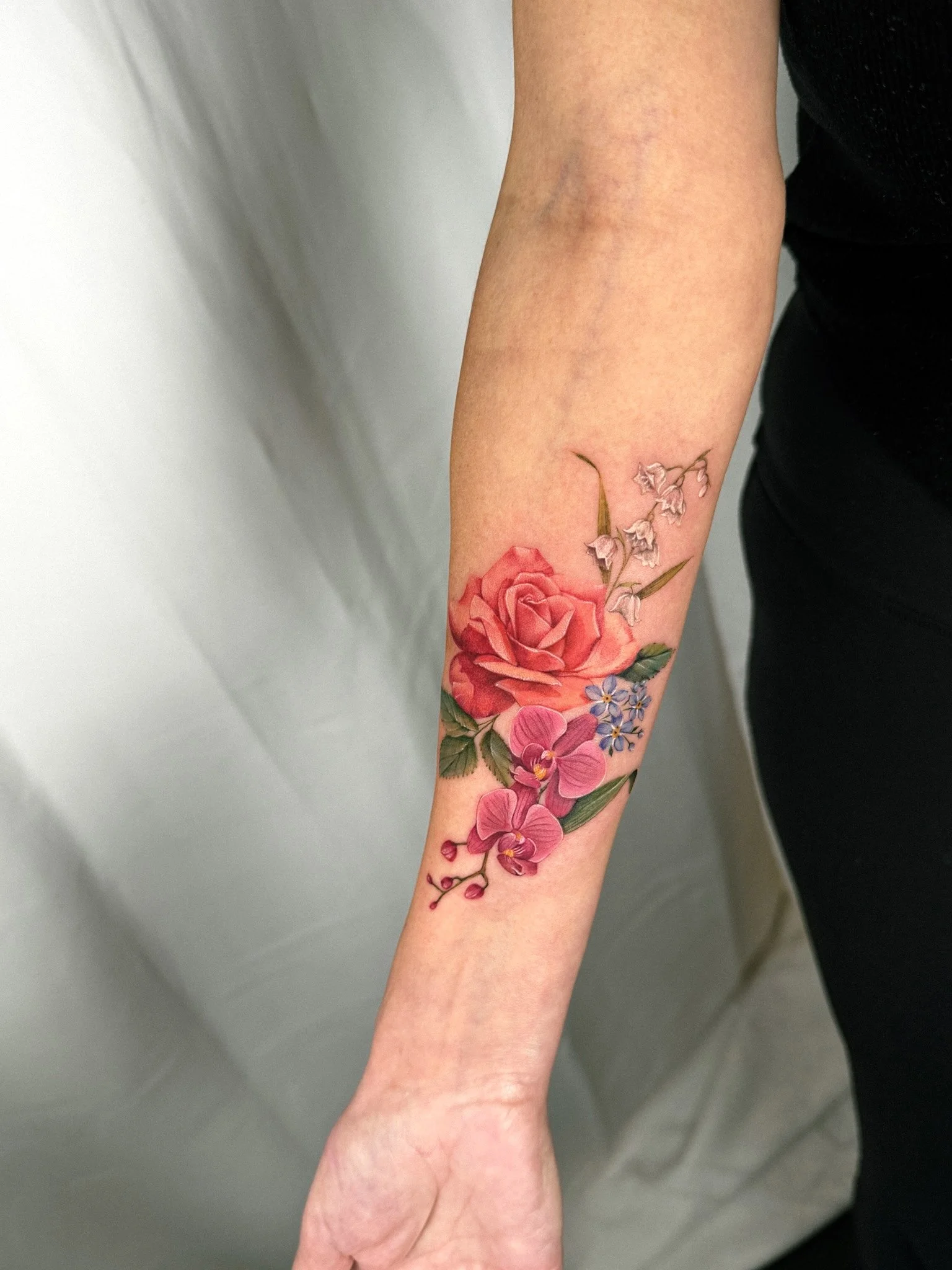 Colorful floral tattoo with a large pink rose, purple orchids, and small white flowers on a person's forearm.