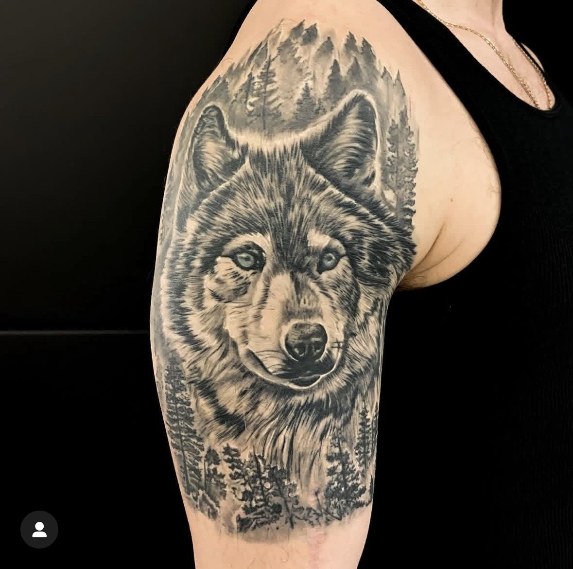 Black and grey tattoo of a wolf's face on a person's upper arm, with a forest scene and mountains in the background.