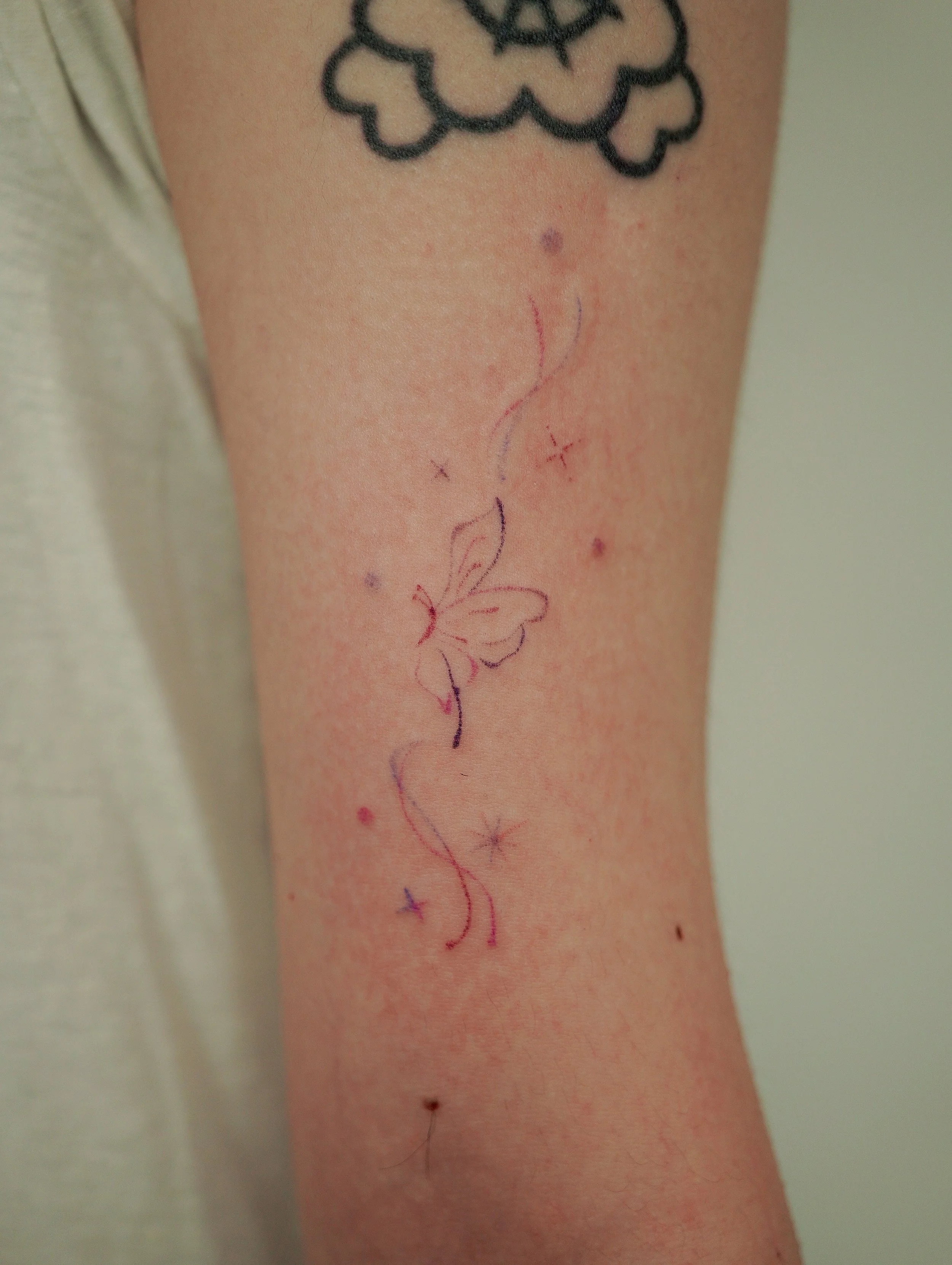 A person's arm with black tattoo art near the shoulder and colorful temporary or henna tattoo of a butterfly with swirling lines and star accents on the lower arm.