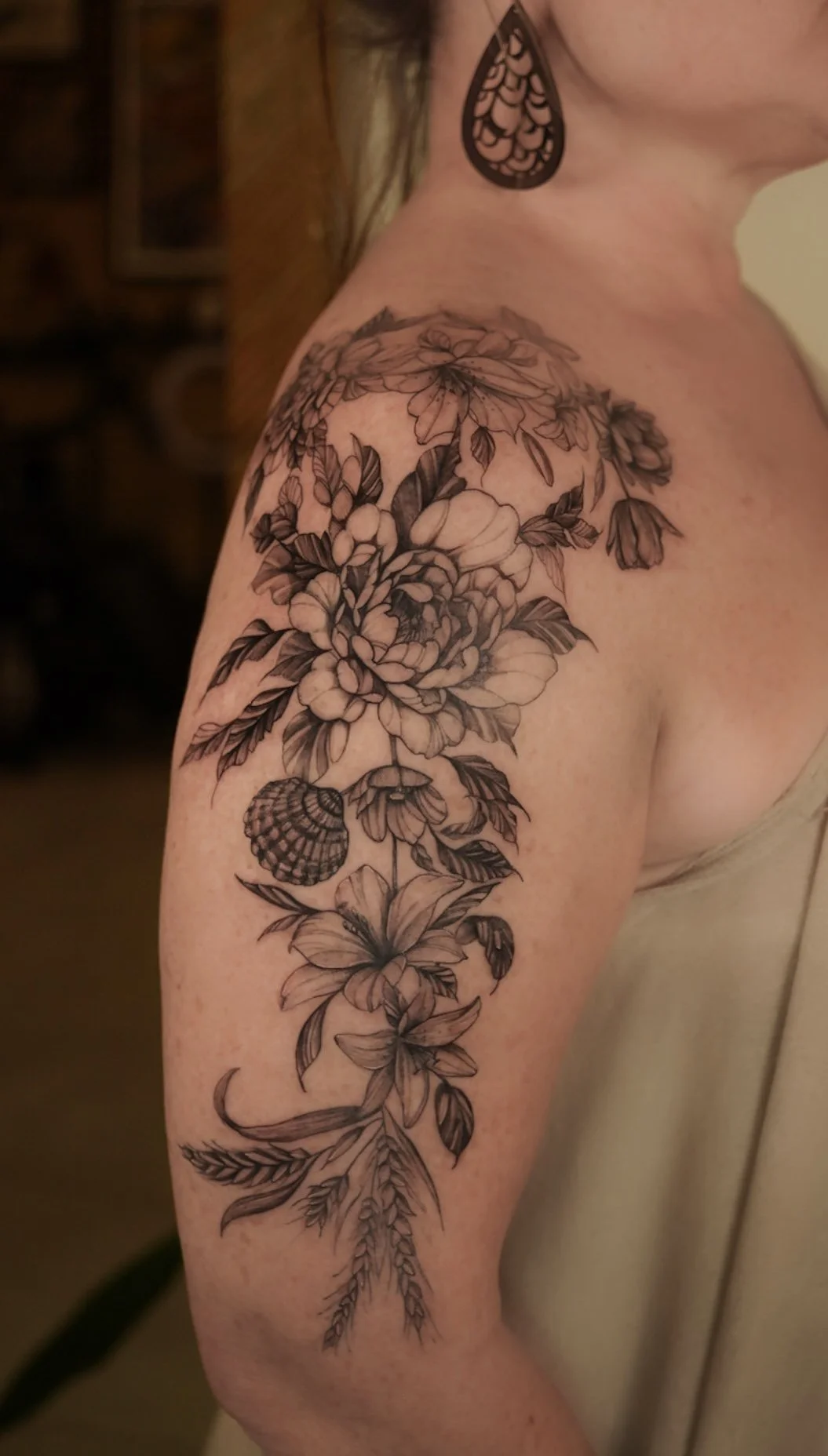 Black and gray floral tattoo on woman's left shoulder and upper arm, featuring detailed flowers, leaves, and a seashell.