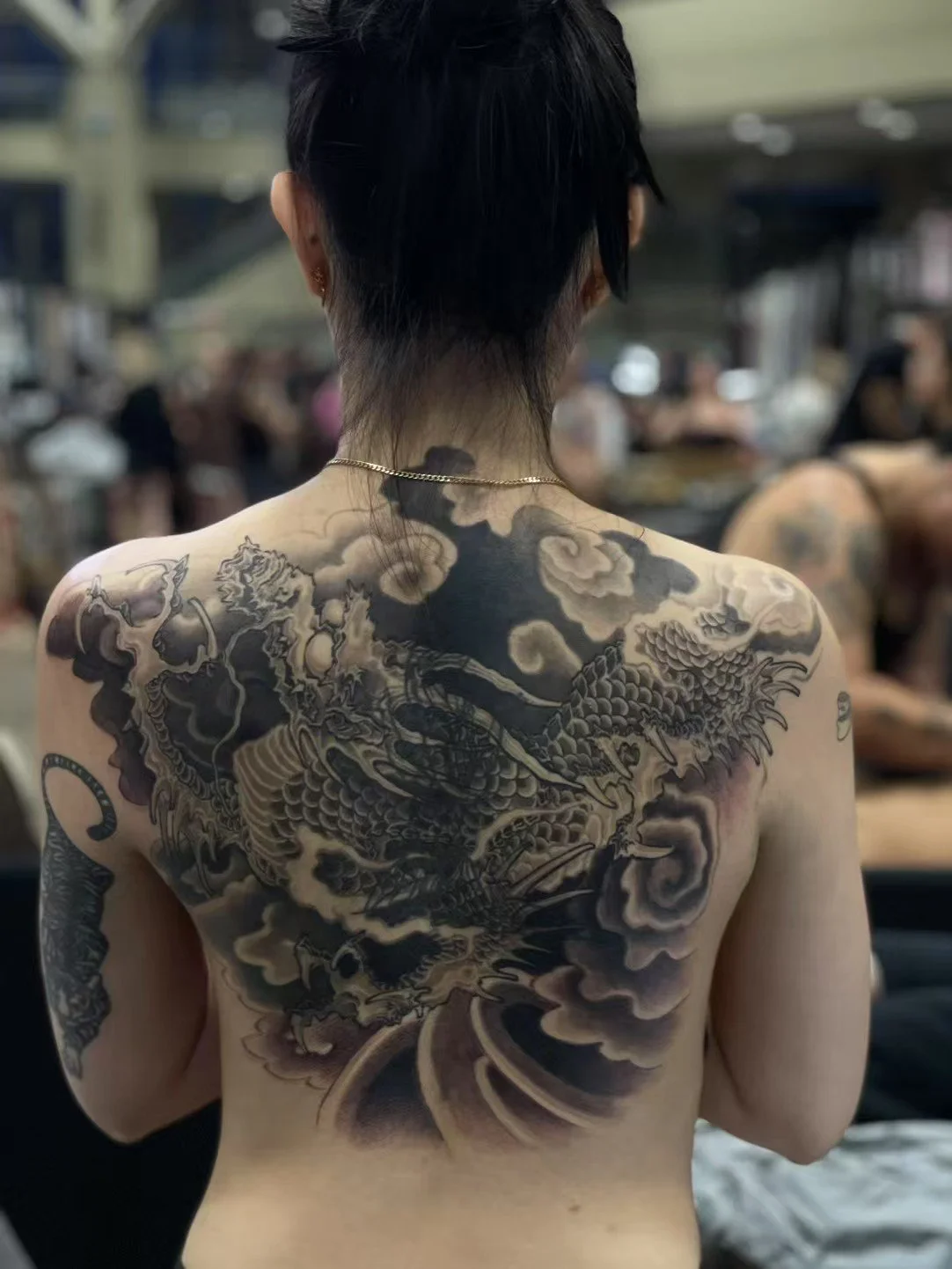 Back view of a person with short black hair displaying a large black and gray dragon tattoo covering the back and shoulders.