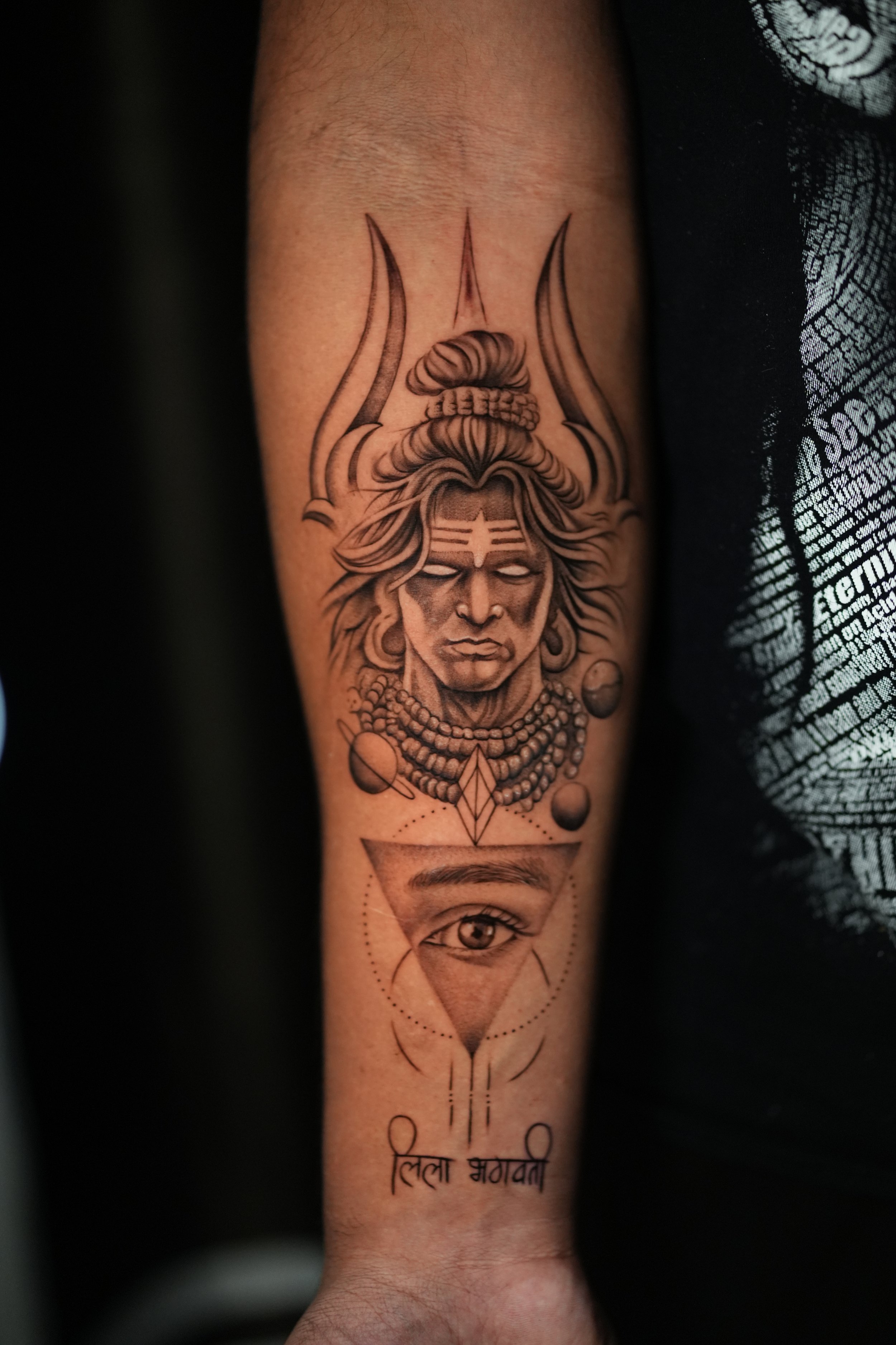 Tattoo featuring a detailed depiction of a revered Hindu goddess with elaborate headgear and multiple necklaces, above an eye within a triangle, with geometric and symbolic elements and Hindi script at the bottom.