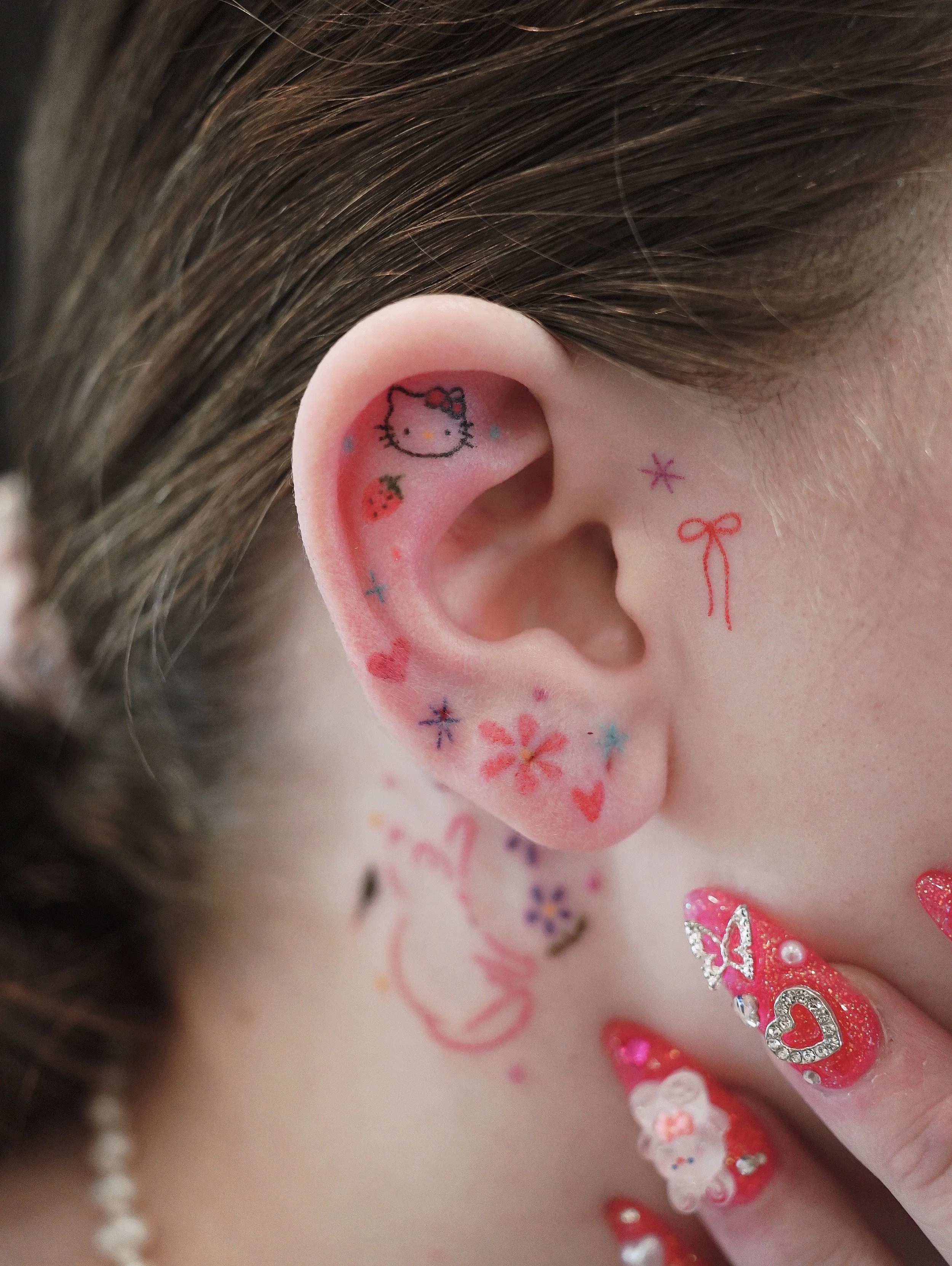 Close-up of a person's ear decorated with various colorful temporary tattoos, including Hello Kitty, hearts, swirls, and floral designs, with a matching floral tattoo on the neck. The person has long, styled hair and decorated nails with pink designs