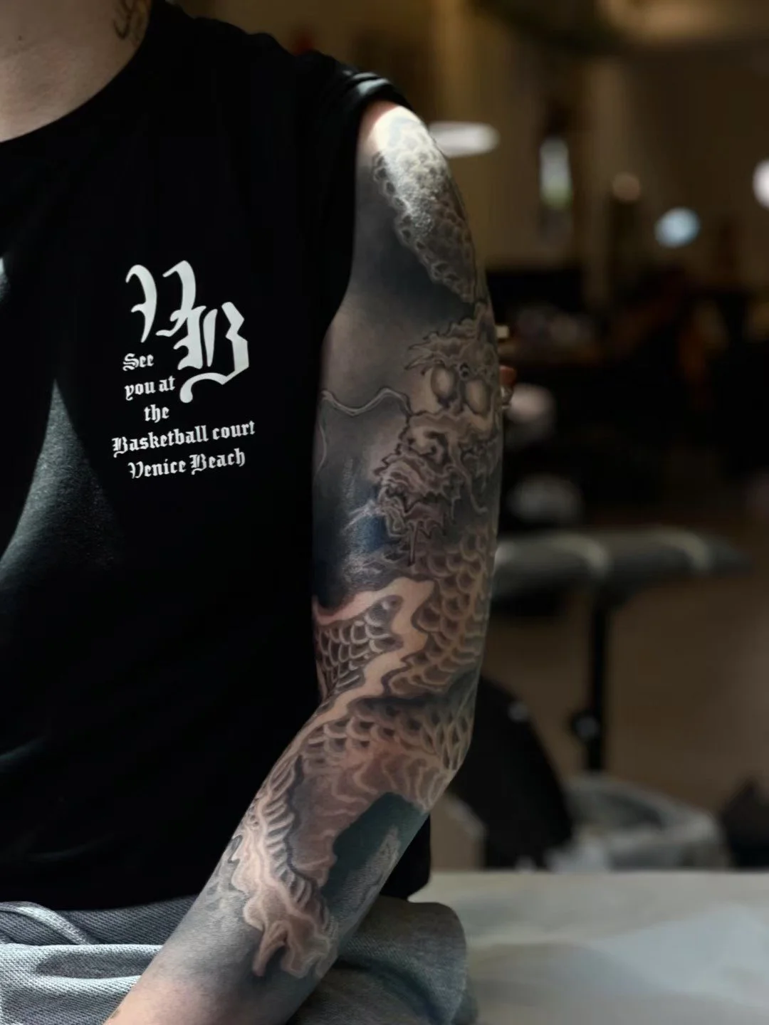 Close-up of a person’s arm with intricate black and gray tattoo artwork featuring clouds, waves, and dragons, wearing a black T-shirt with white text and a gothic-style logo, in a dimly lit indoor environment.