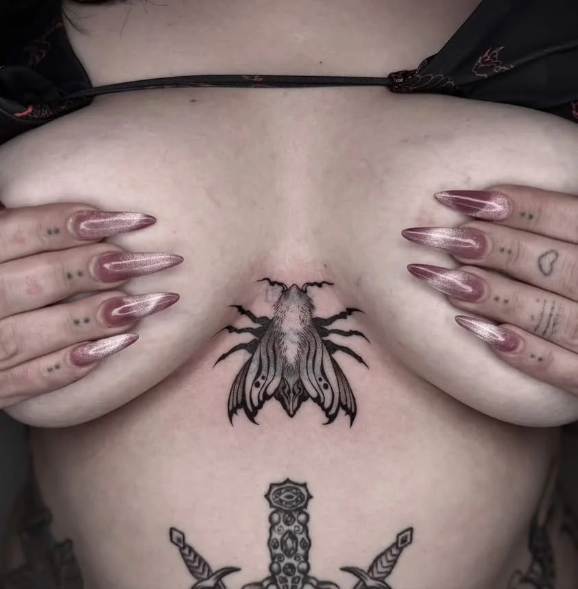 Close-up of a person's chest with a large moth tattoo in the center and additional tattoos on both hands and fingers, including small dots and hearts.