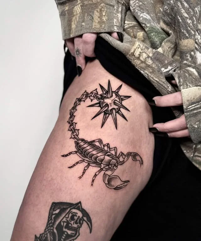 Tattoo of a scorpion and a spiked mace on a person's thigh, with a partially visible skull tattoo below.