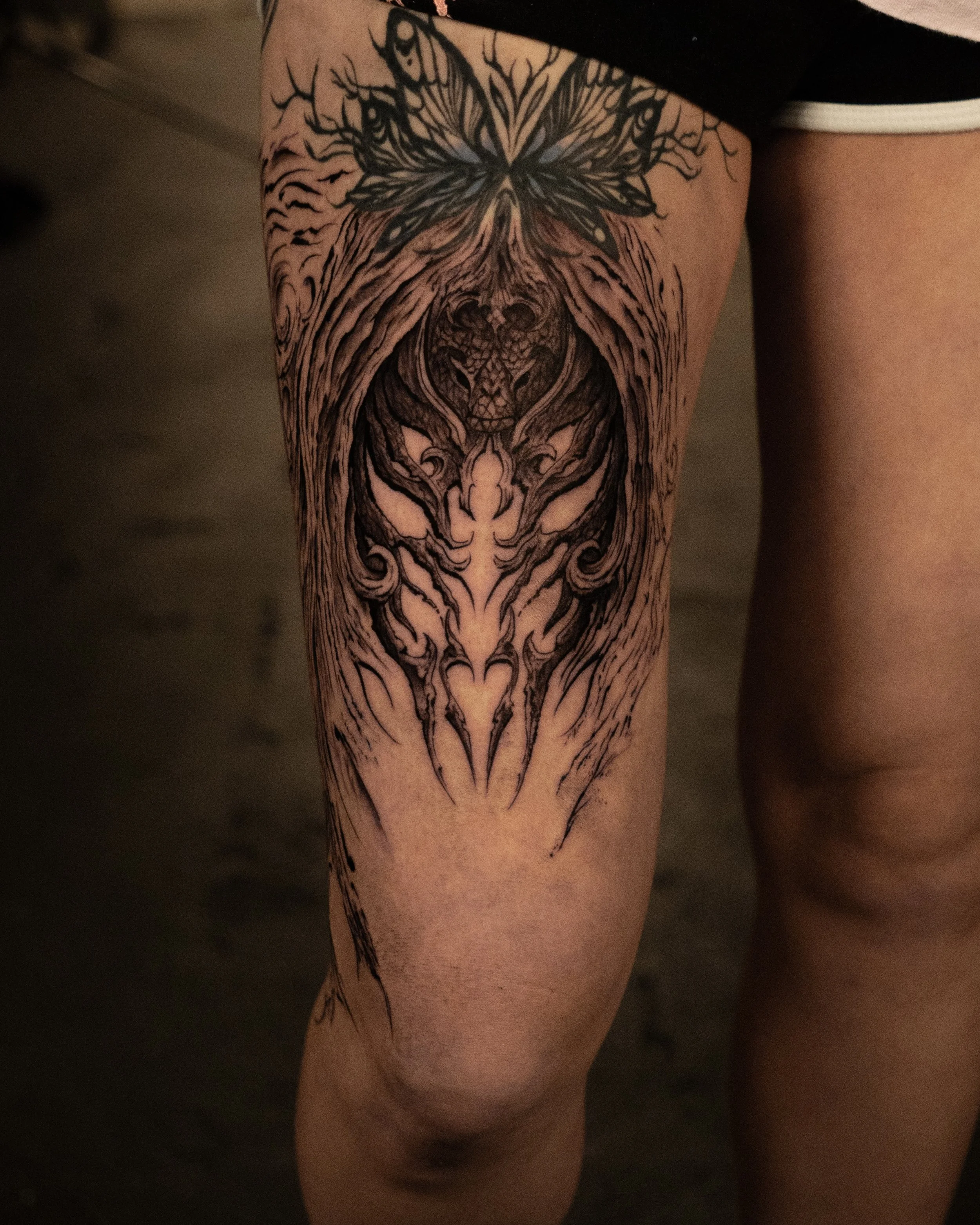 Tattoo of a dragon's face with intricate lines on a person's thigh, with a butterfly near the top.