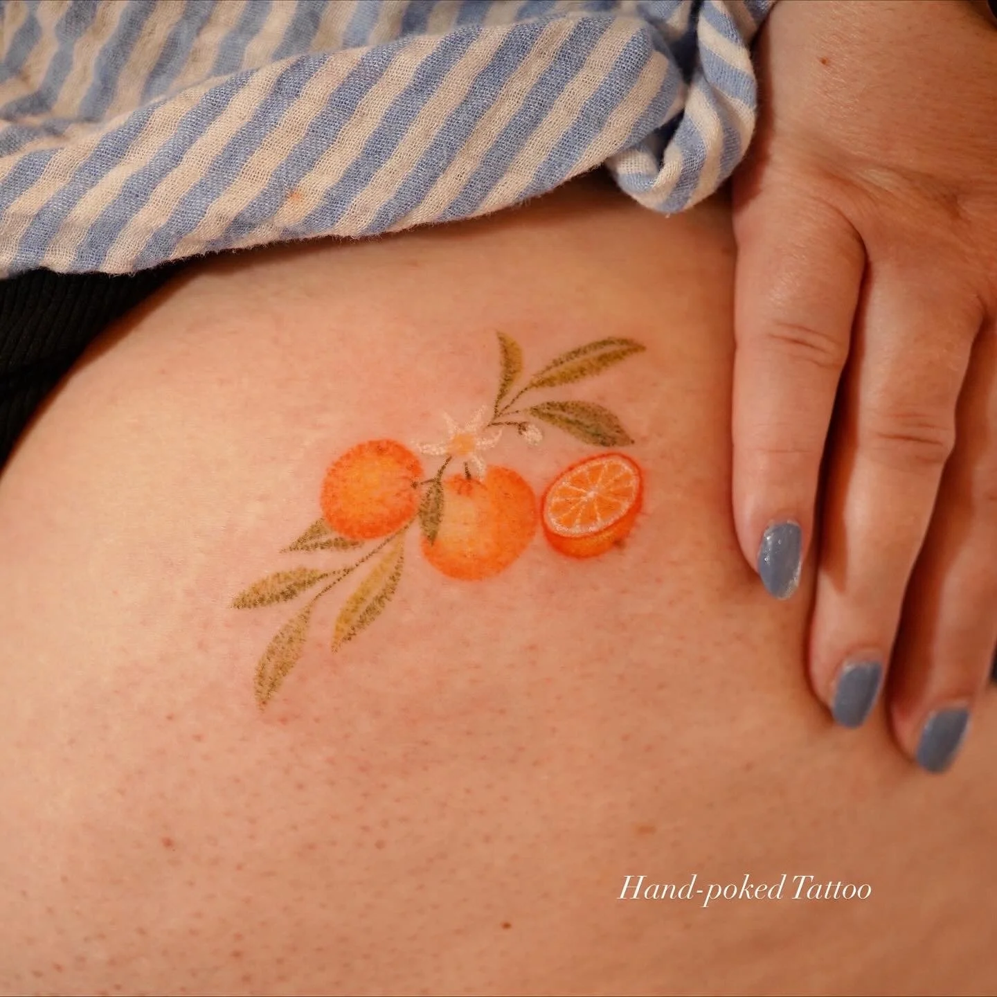 Tattoo of oranges and orange leaves on skin with a person's hand nearby. Text reads 'Hand-poked Tattoo'.