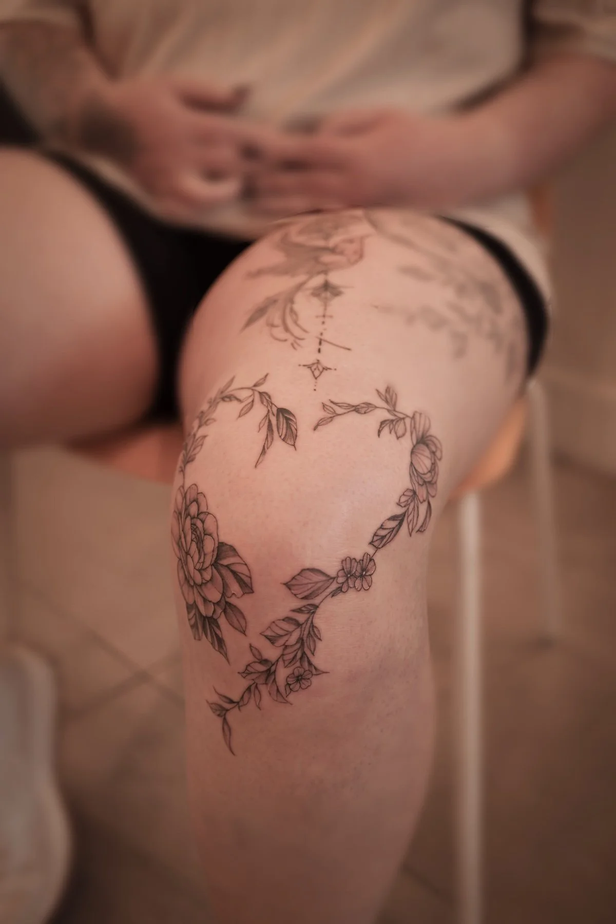 A person's thigh with a floral heart-shaped tattoo outline, featuring detailed flowers and leaves.