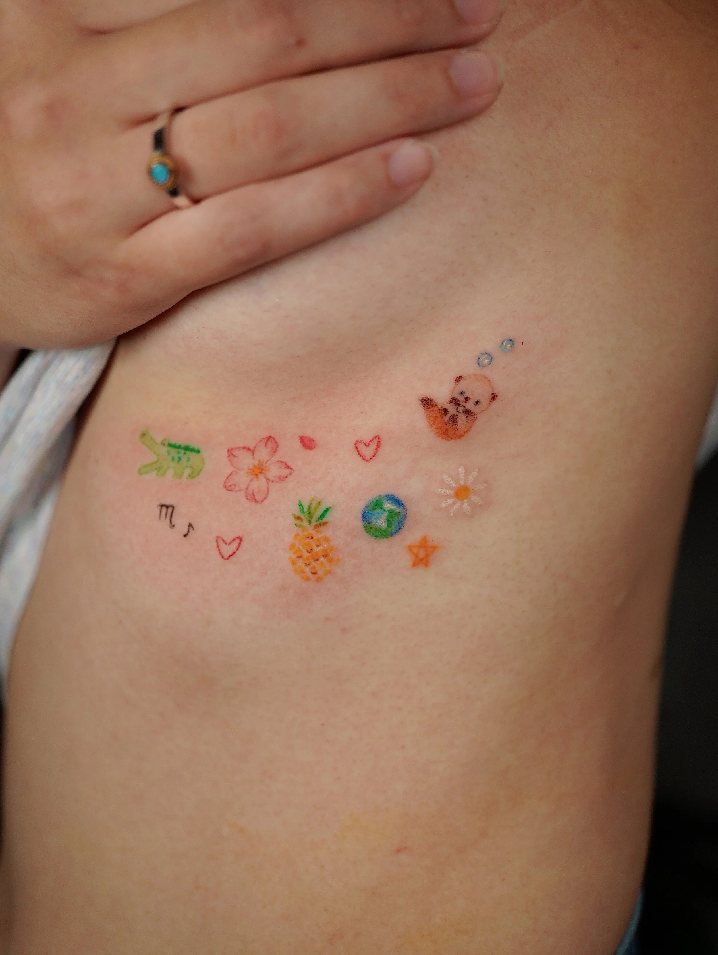 A person's chest with colorful temporary tattoos of various small images, including a crocodile, cherry blossom, pineapple, the Earth, a star, a daisy, a bear, and hearts, with a hand partially covering the upper part of the chest.