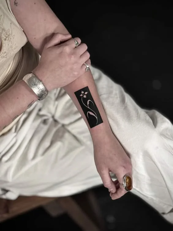 A person with a wrist tattoo of stylized flowers in black ink on their arm, wearing a silver bracelet and rings, dressed in light-colored clothing, sitting with their arm resting on their lap.