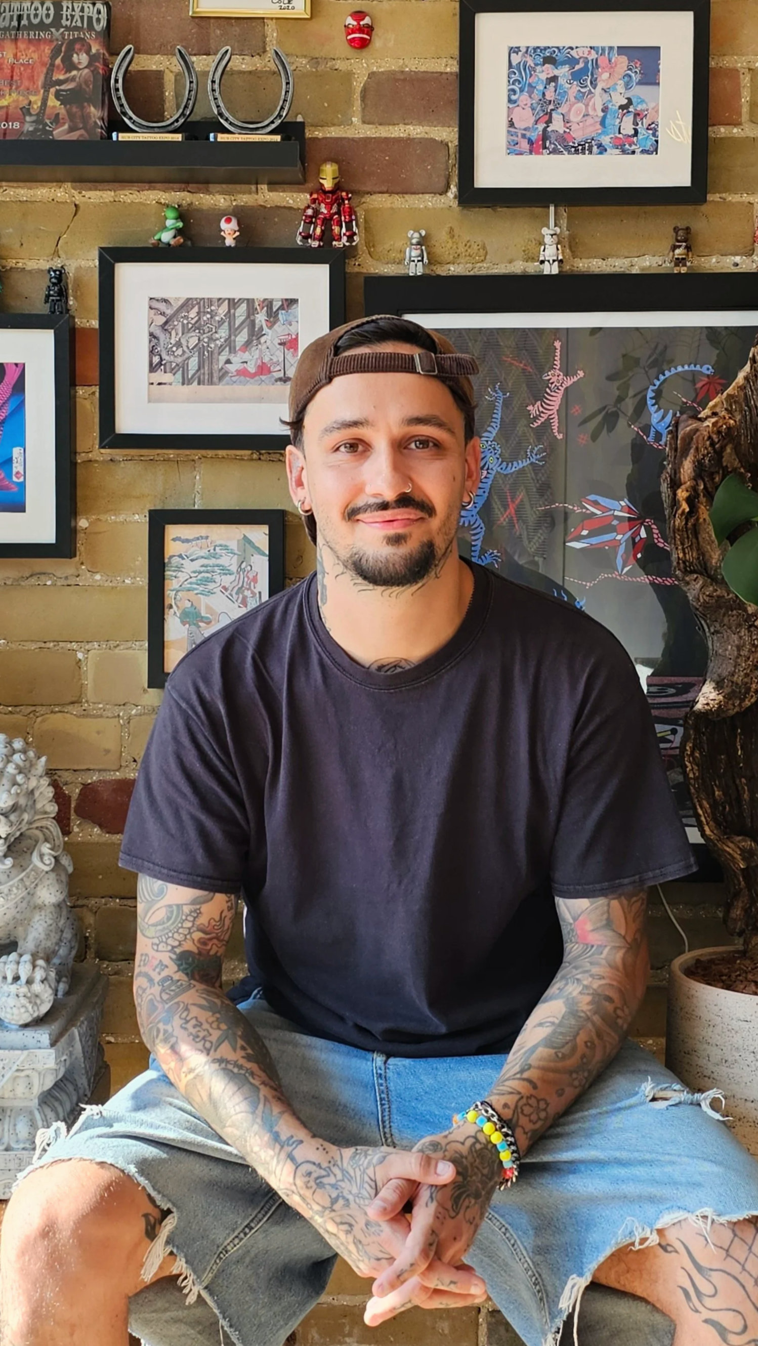 Young man with tattoos, wearing a dark t-shirt and light denim shorts, sits with his hands clasped in front of a brick wall decorated with framed artwork, small figurines, and decorative objects.