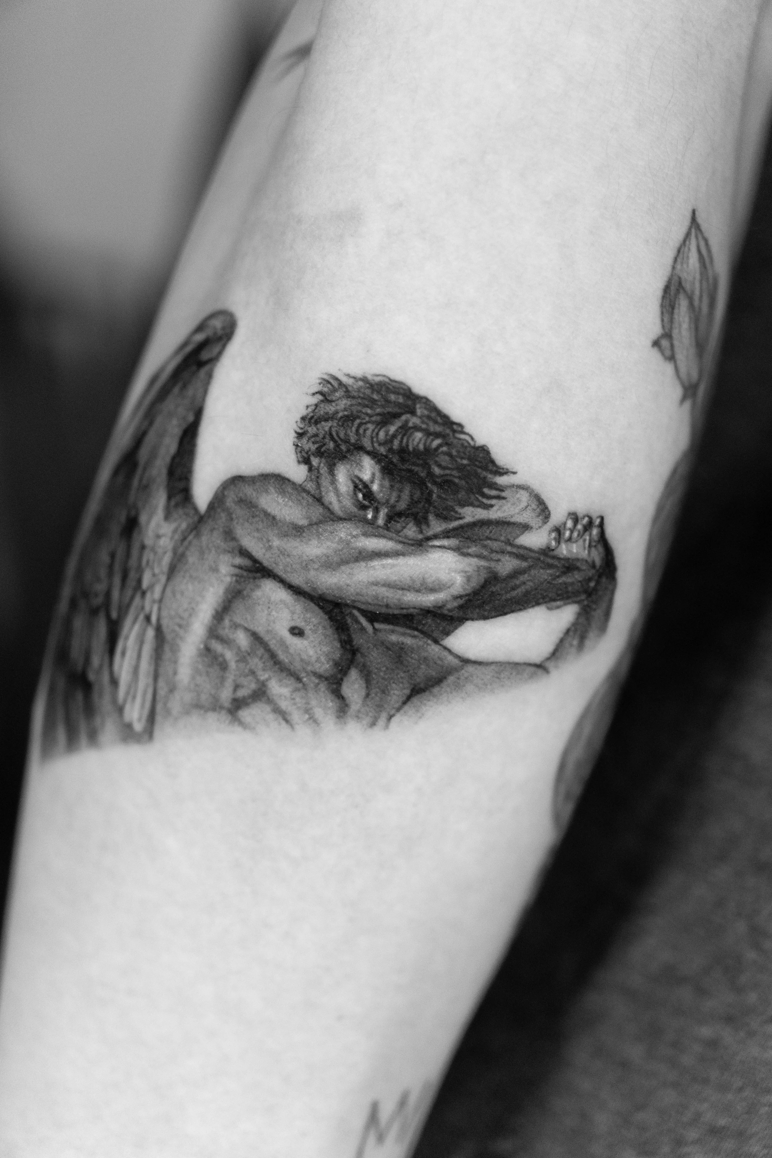 Black and gray tattoo of a winged figure with curly hair, resting her chin on her crossed arms.