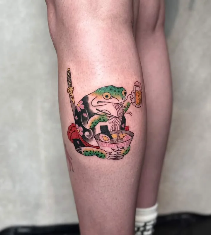 Colorful tattoo on thigh of a frog eating noodles with chopsticks, holding a beer mug, and sitting in a bowl with sushi and chopsticks.