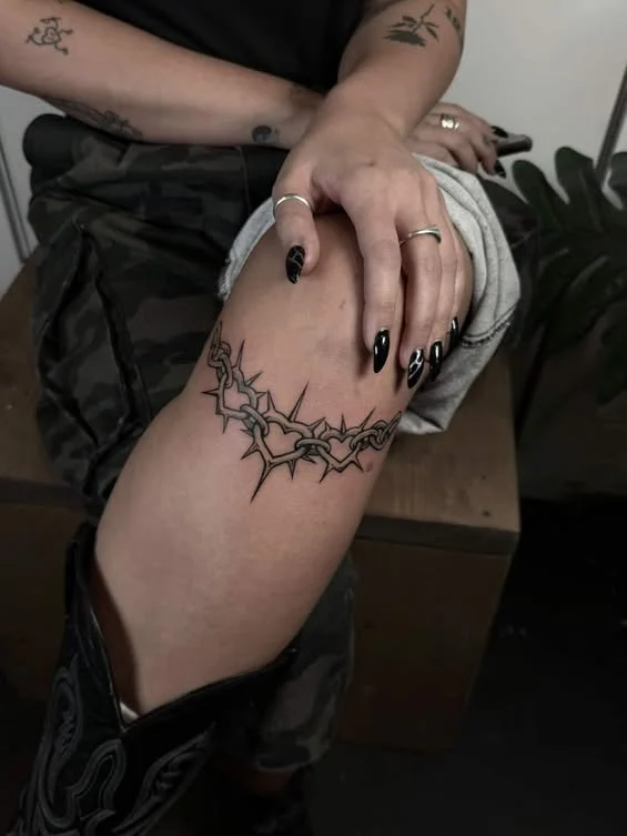 Person with black painted nails and tattoos on arm, holding their thigh to reveal a large, intricate black tattoo resembling a thorny vine or barbed wire.