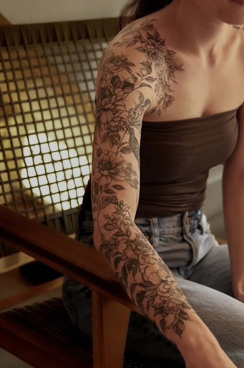 A woman with a large floral tattoo on her arm, wearing a strapless top and jeans, sitting on a wooden chair in a cozy, warmly lit room.