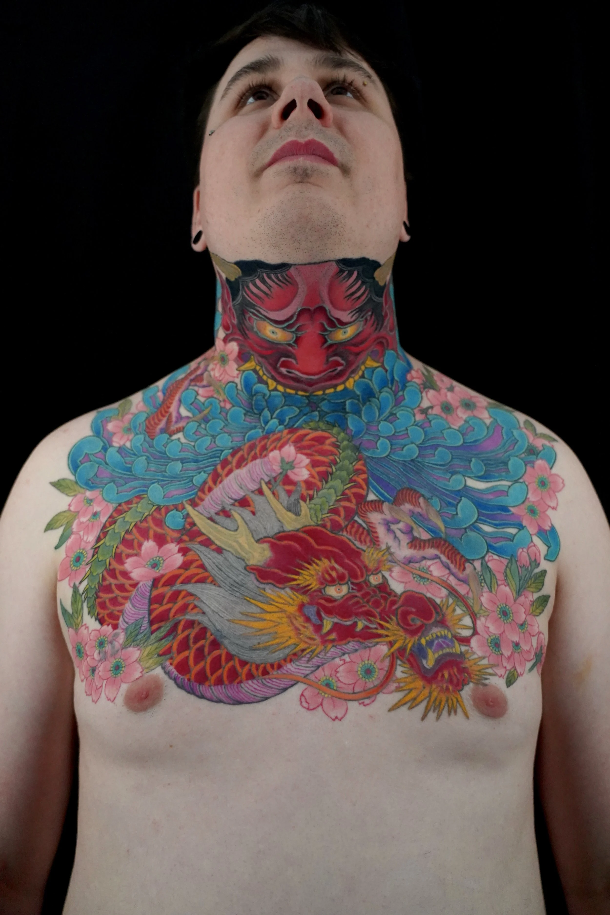 A person with full body tattoos of traditional Japanese-style art, including a prominent face of a figure with an intense expression, holding swords, with intricate details and floral patterns on the arms and torso.