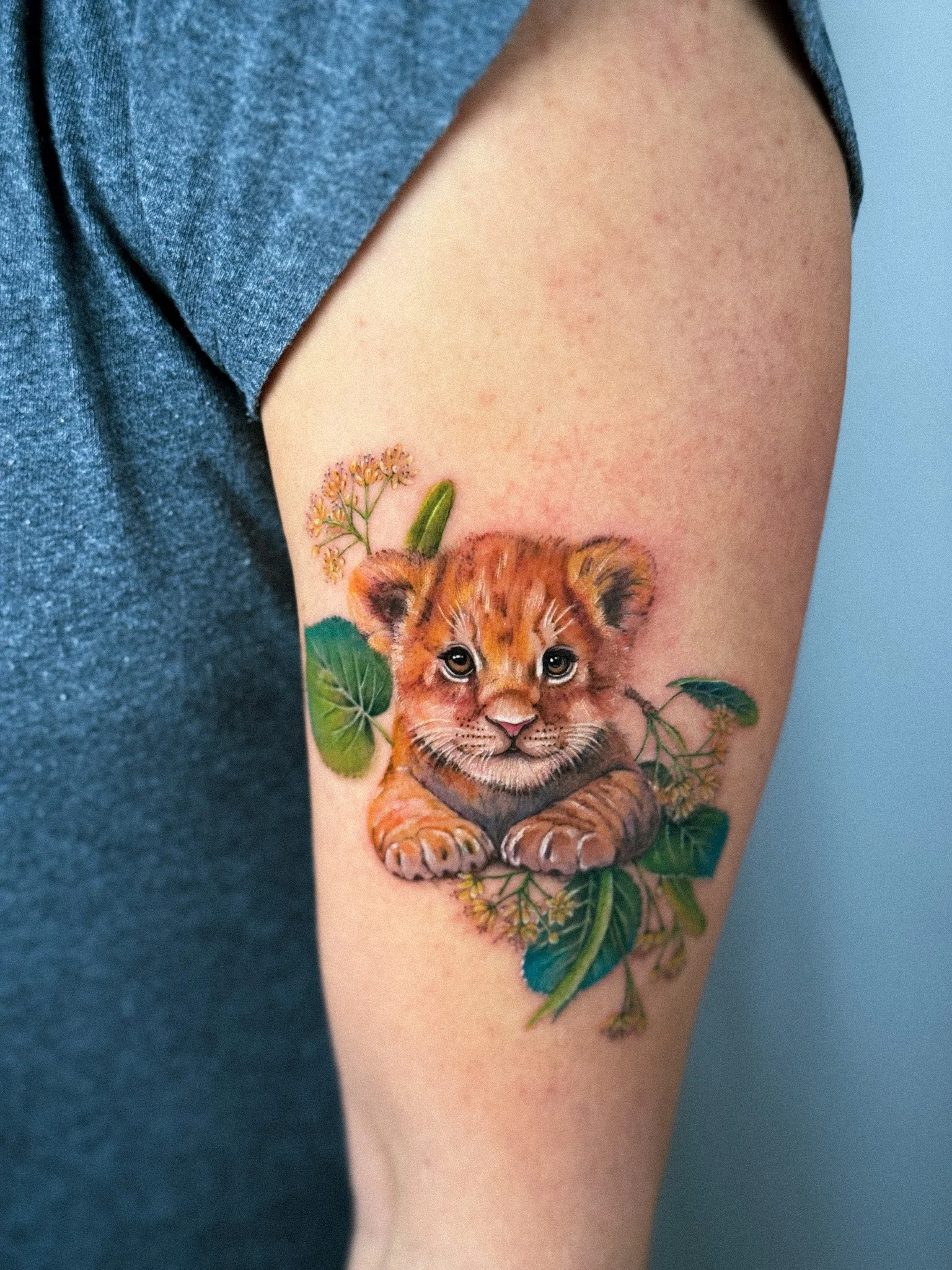 Colorful tattoo of a baby lion cub surrounded by green leaves and small pink flowers on a person's upper arm.