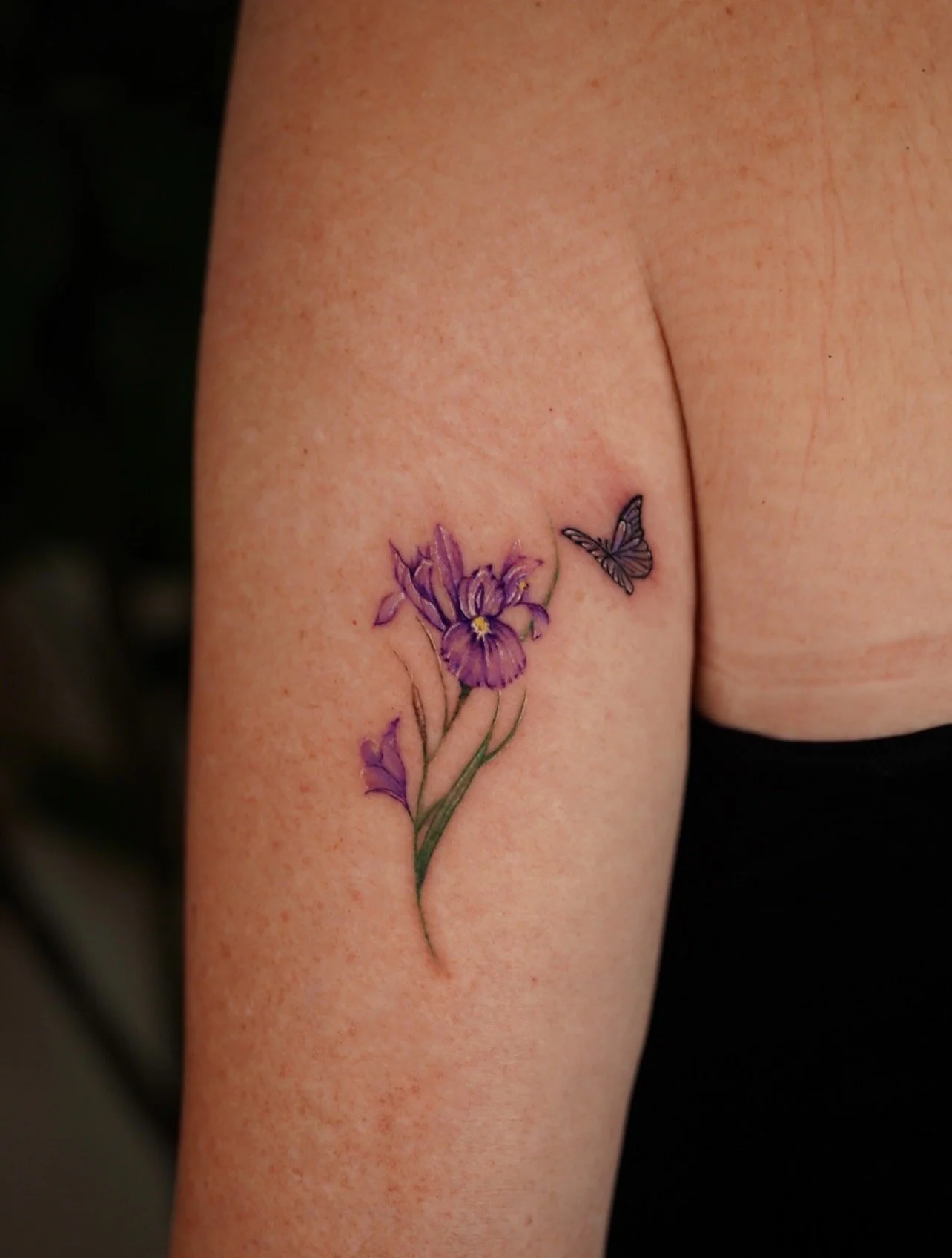 Tattoo of a purple iris flower with green stem and leaves, and a small gray and purple butterfly on the upper right.