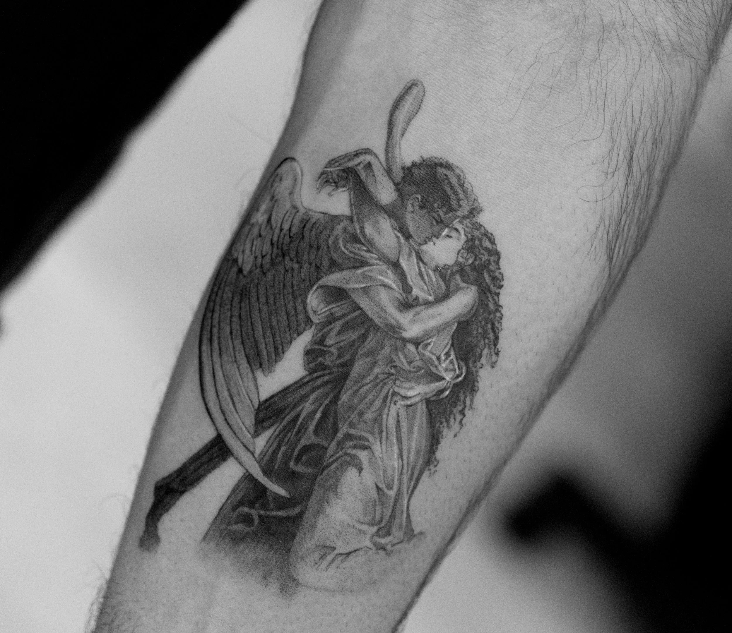 A black and gray tattoo of a angel with wings holding a woman with curly hair in an embrace on someone's arm.