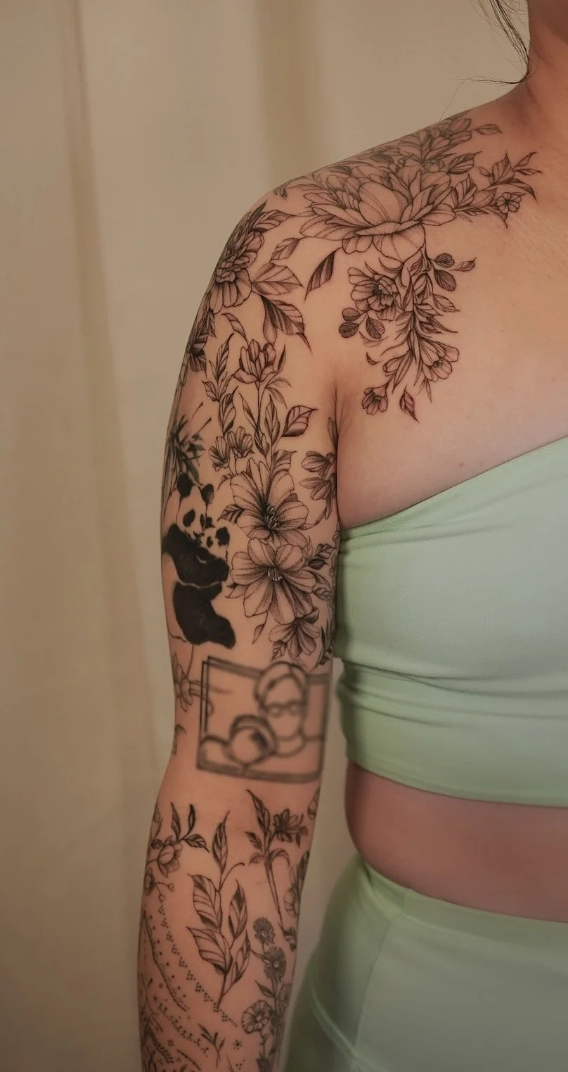 Close-up of a person with a light green tube top and extensive black and gray floral tattoos on the shoulder and arm.